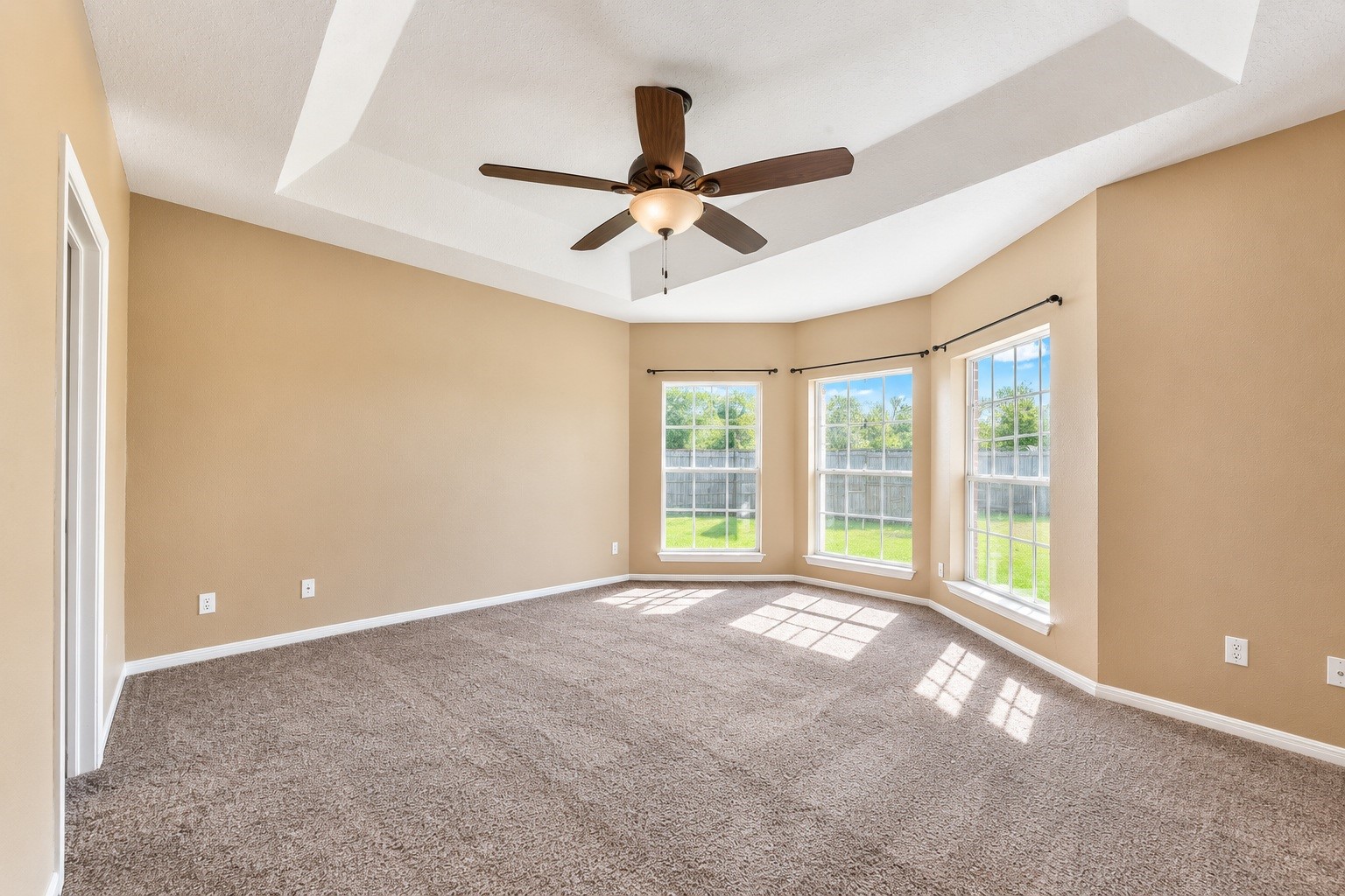 Spacious master bedroom with a tray ceiling, modern ceiling fan, and large bay windows offering abundant natural light and serene backyard views.