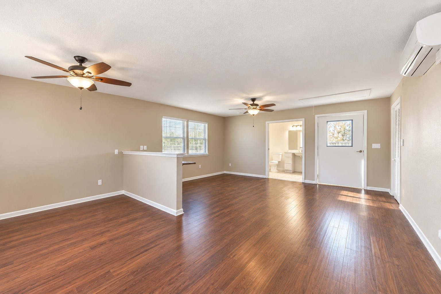 Spacious detached living area with rich wood-look flooring, neutral walls, and ceiling fans, leading to a bright bathroom and adjacent to a cozy nook with natural light.