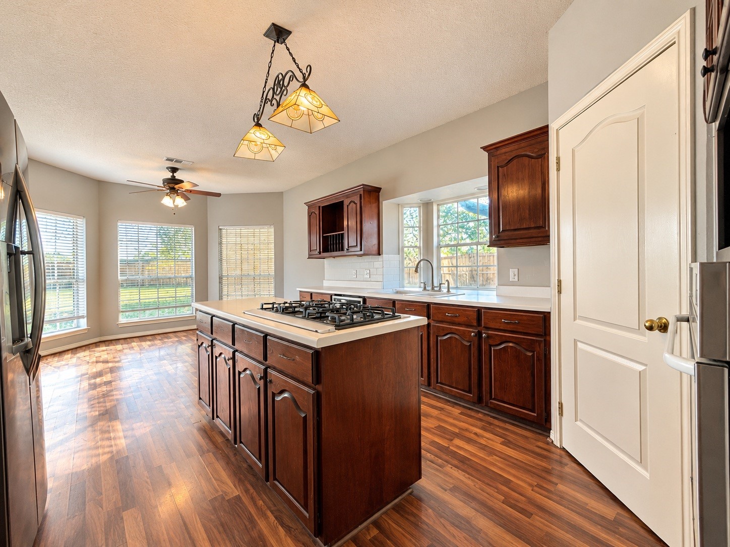 Spacious kitchen featuring rich wooden cabinetry, a central island with a gas cooktop, and large windows offering abundant natural light and views of the backyard.