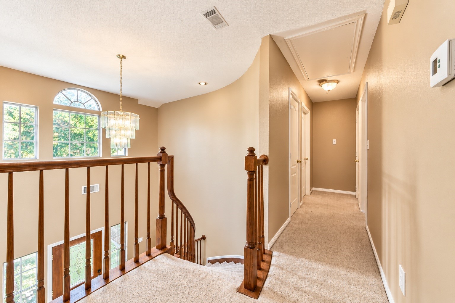 Bright and inviting upstairs hallway with a charming curved staircase, elegant chandelier, and ample natural light from large windows.