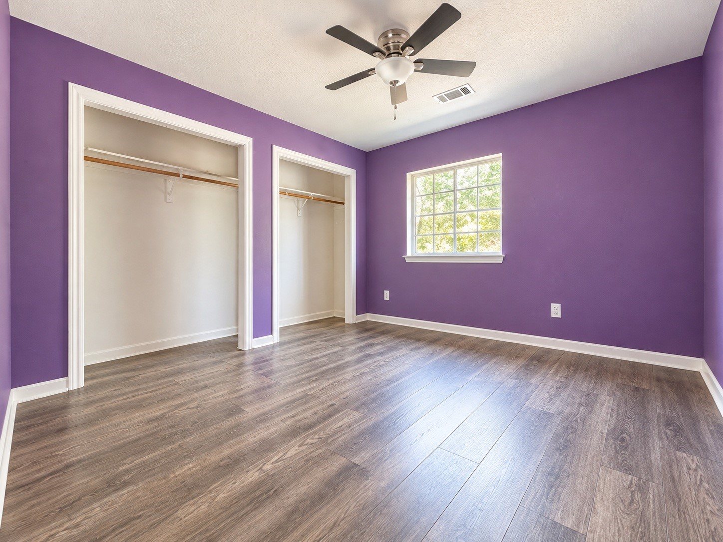 Bright bedroom with vibrant purple walls, dual closets, and warm wood-look flooring, complemented by a ceiling fan and a large window for natural light.