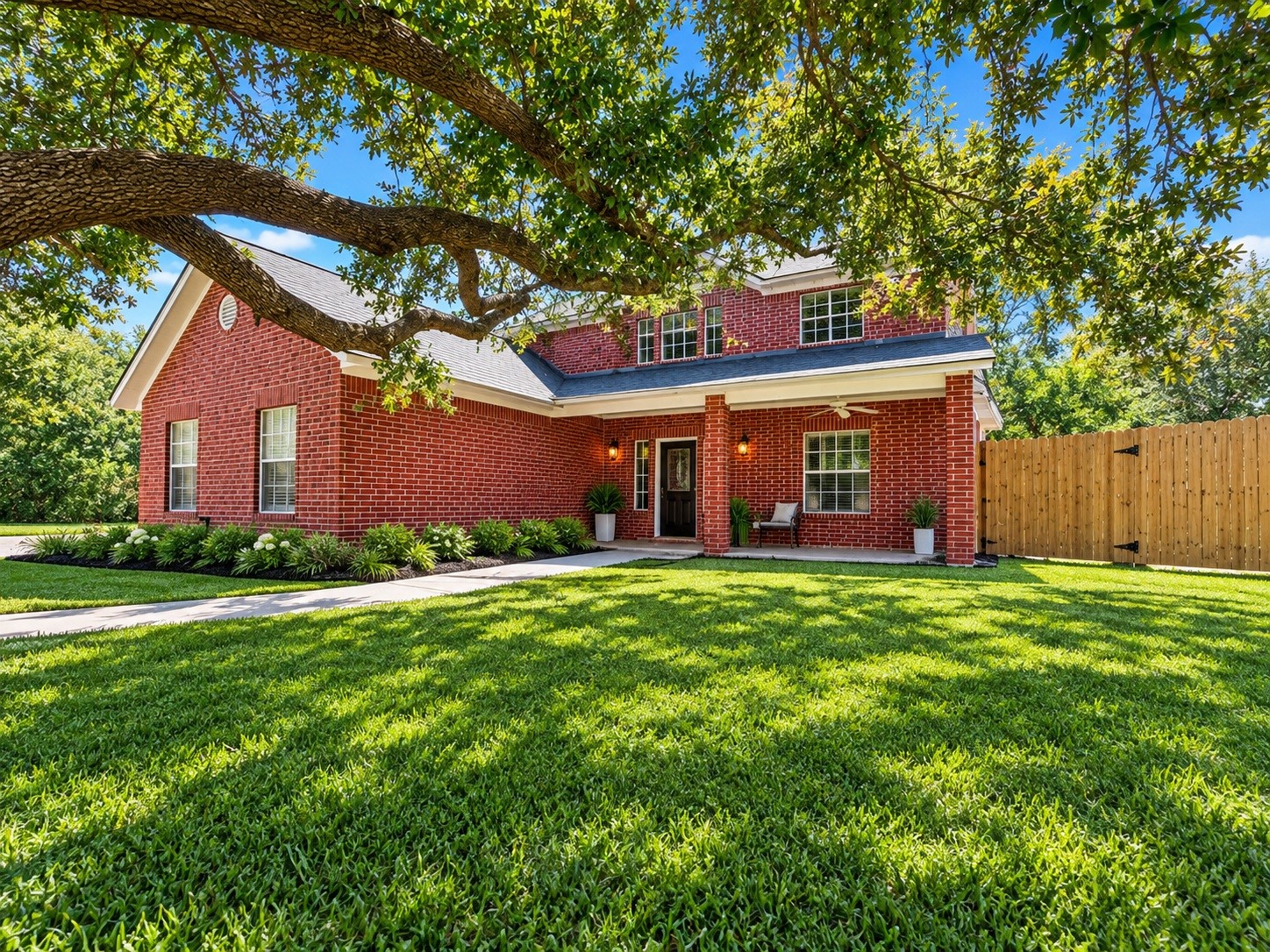 Charming red brick home with a welcoming front porch, surrounded by lush greenery and mature trees, offering a serene and picturesque setting. Landscaping virtually added for appeal.