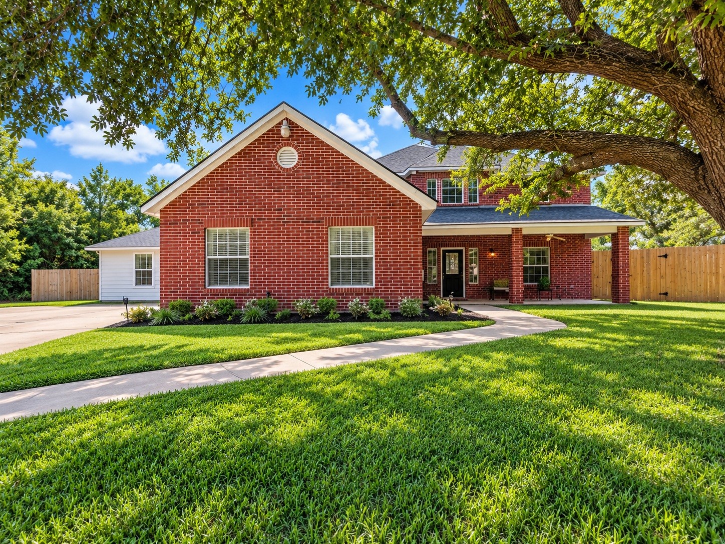 Charming red brick home with a welcoming front porch, nestled under a mature shade tree, features a well-manicured lawn and a spacious driveway. Landscaping virtually added for appeal.