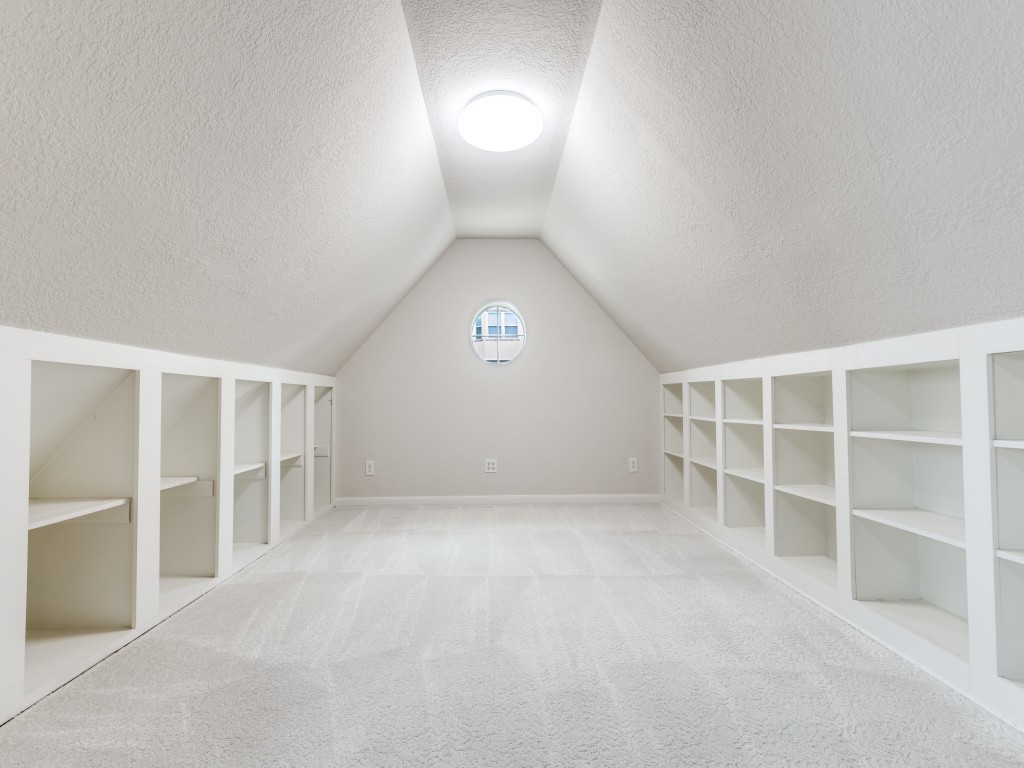 This photo showcases the nook off the 2nd bedroom space. A barn door separates it from the main room. It has a sloped ceiling, built-in shelving along both sides for ample storage. The room is well-lit by a circular window at the end and a ceiling light, offering a clean, carpeted floor.