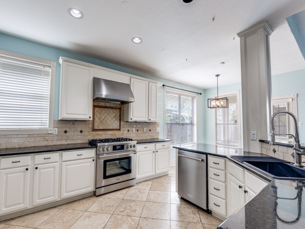Bright and spacious kitchen with modern stainless steel appliances, white cabinetry, and black granite countertops. It features a gas stove, tiled backsplash, and large windows for natural light. The open layout includes a dining area with a stylish pendant light.