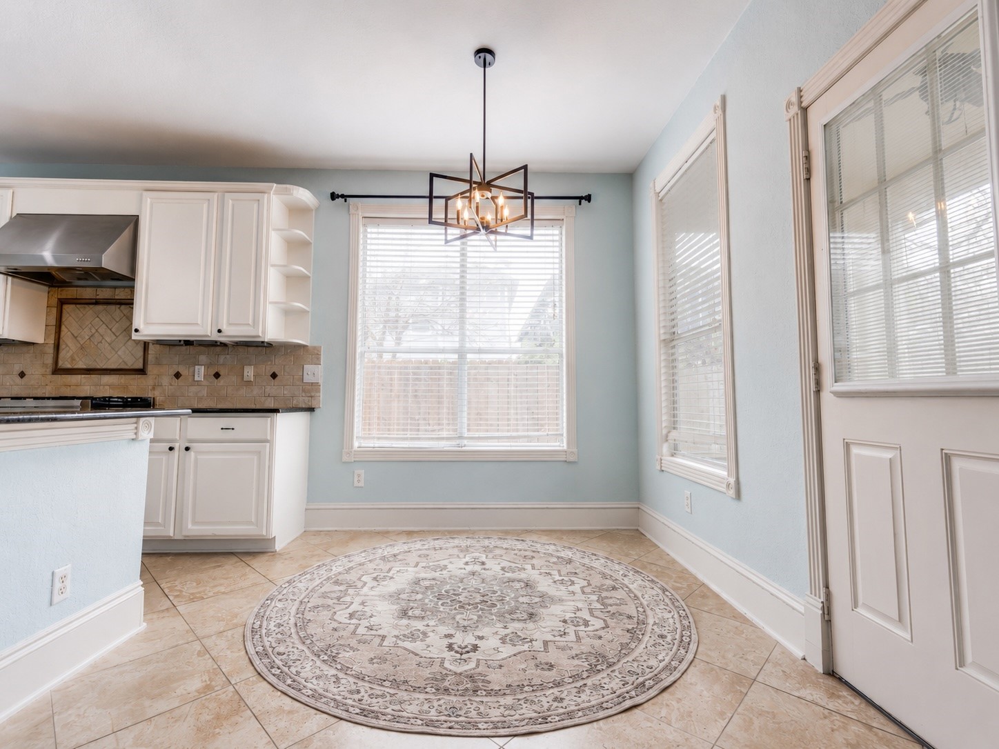 Bright kitchen area with large windows, modern light fixture.Features tiled flooring, white cabinets, and a glass-panel door, creating an inviting and airy atmosphere.