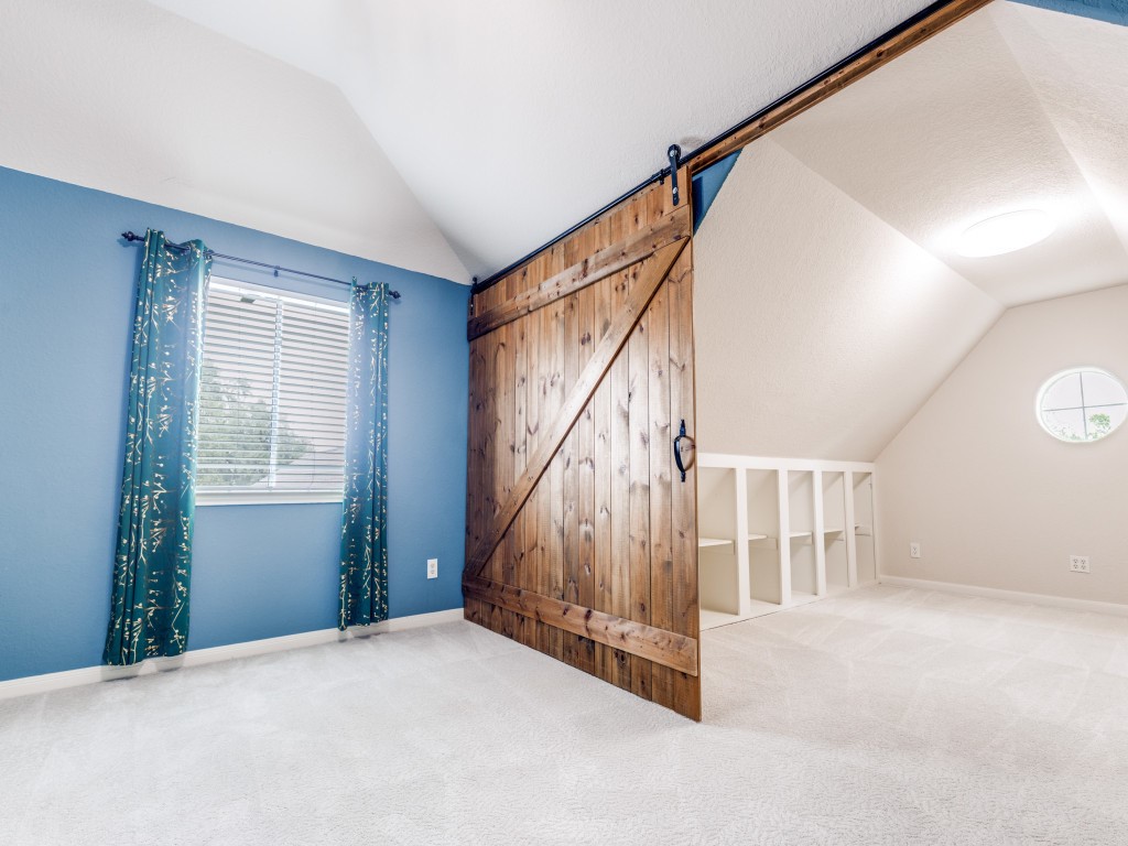 This room features a vaulted ceiling, a rustic sliding barn door, and a cozy nook with built-in shelving. The space is brightened by a window with decorative curtains, offering a charming and functional area which might make a perfect workout room or nursery.