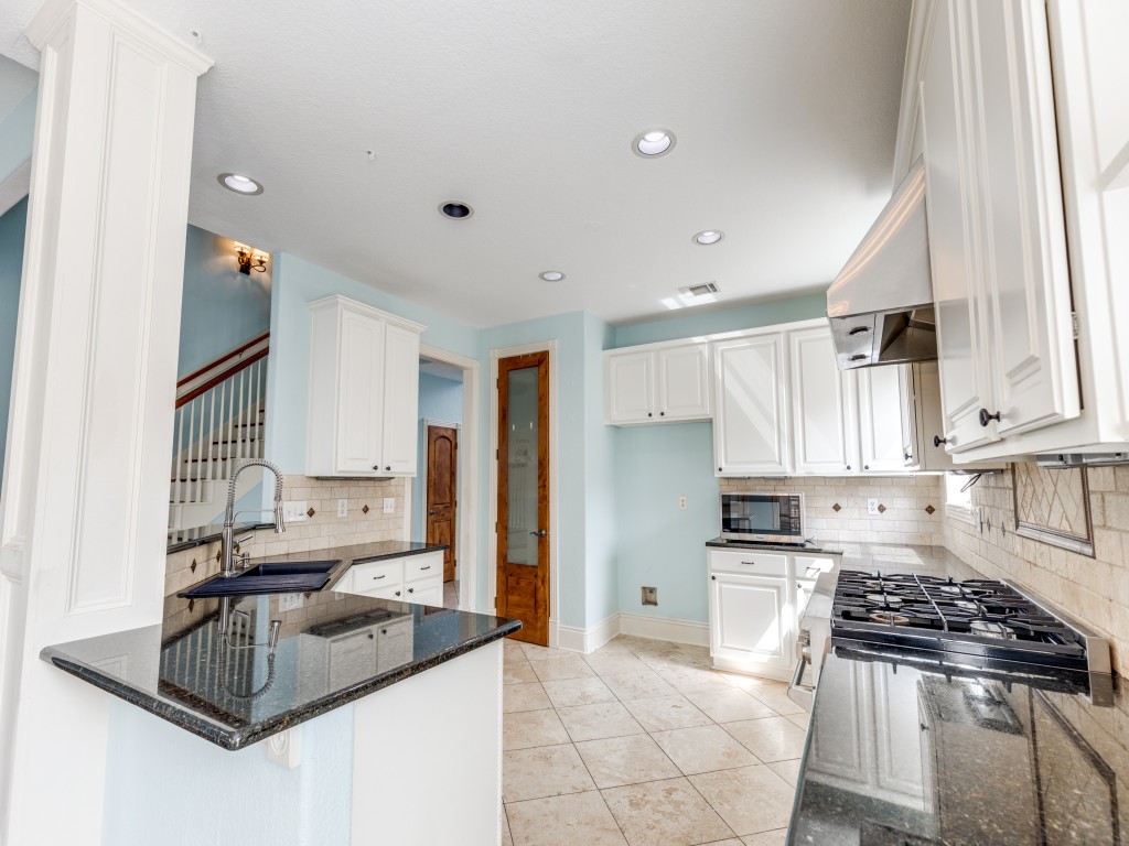 This kitchen features modern white cabinetry, a gas stove, and granite countertops. It has a tiled backsplash, recessed lighting, and a bright, airy feel with light blue walls. The layout includes a breakfast bar and a view of the staircase, offering a spacious and inviting atmosphere.