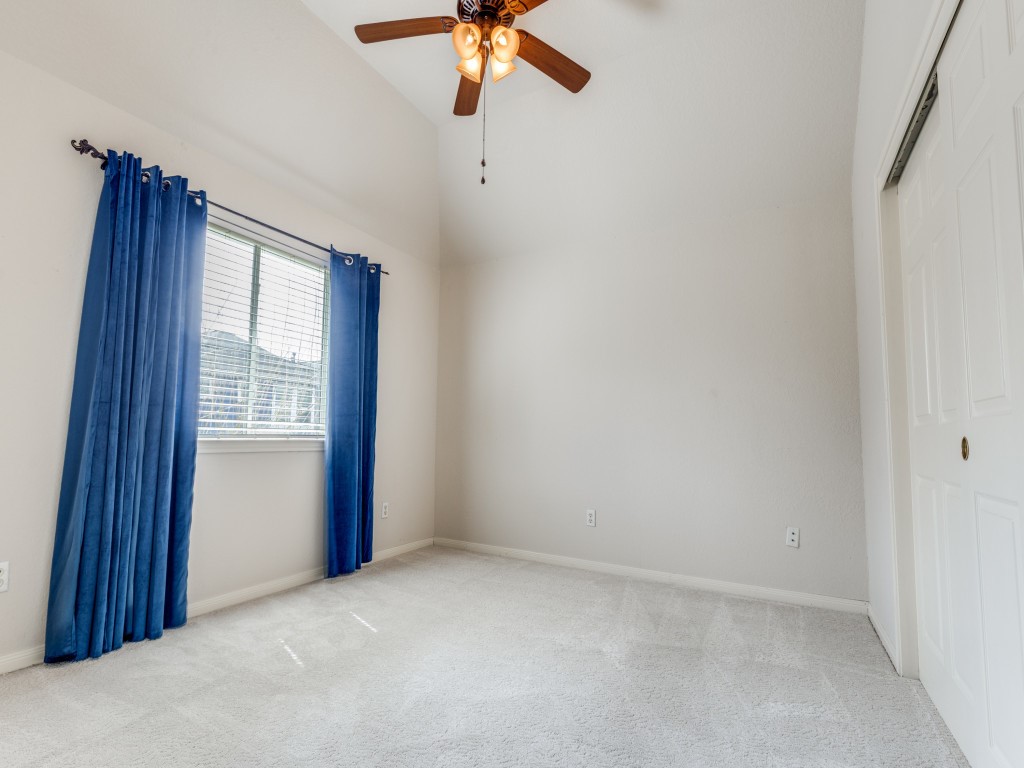 Second floor bedroom with vaulted ceiling, ceiling fan, and large window adorned with blue curtains. Features plush carpeting and a sliding door closet.