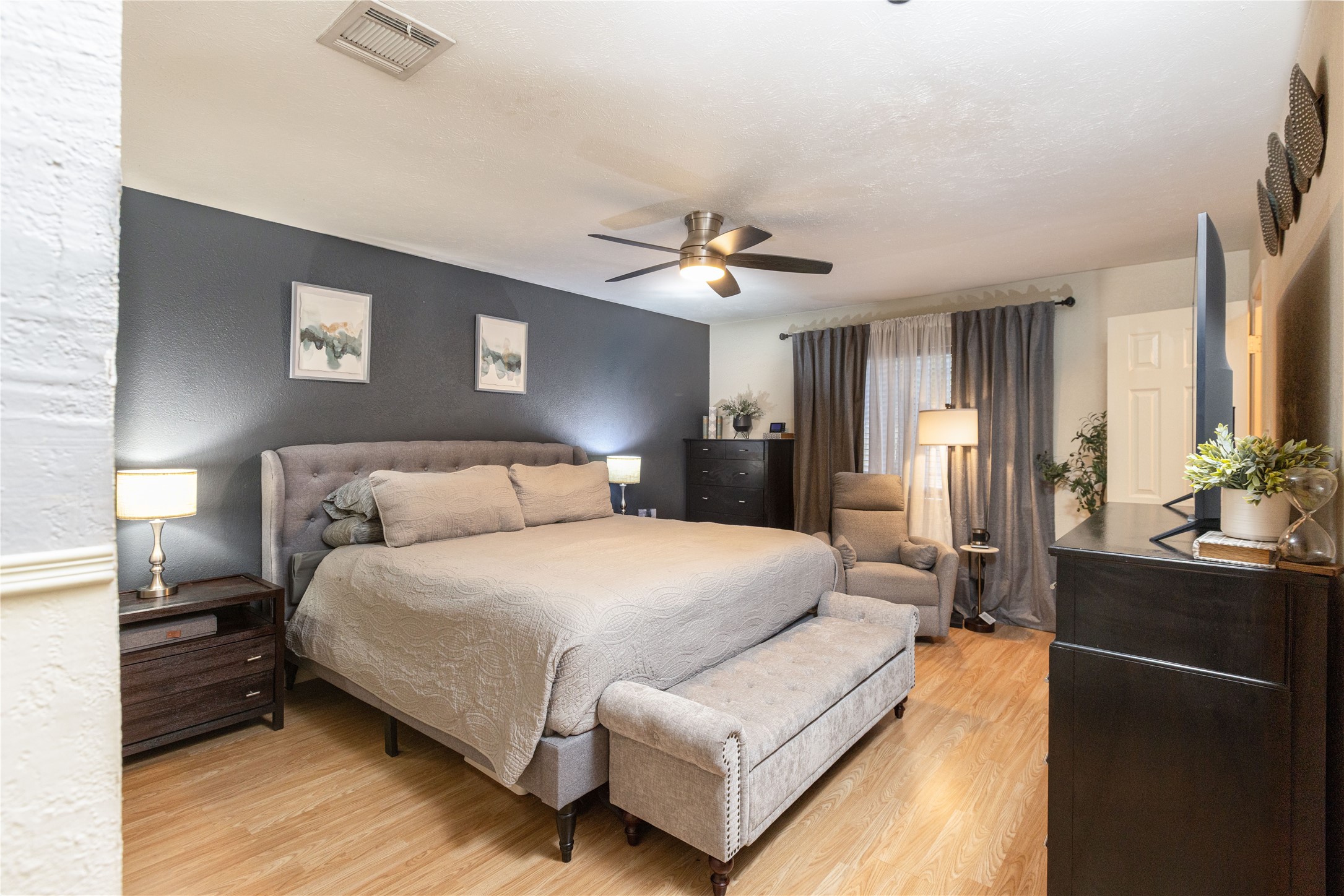 Spacious bedroom featuring a cozy bed, elegant dark accent wall, and modern ceiling fan, complemented by stylish furniture and warm wood flooring.