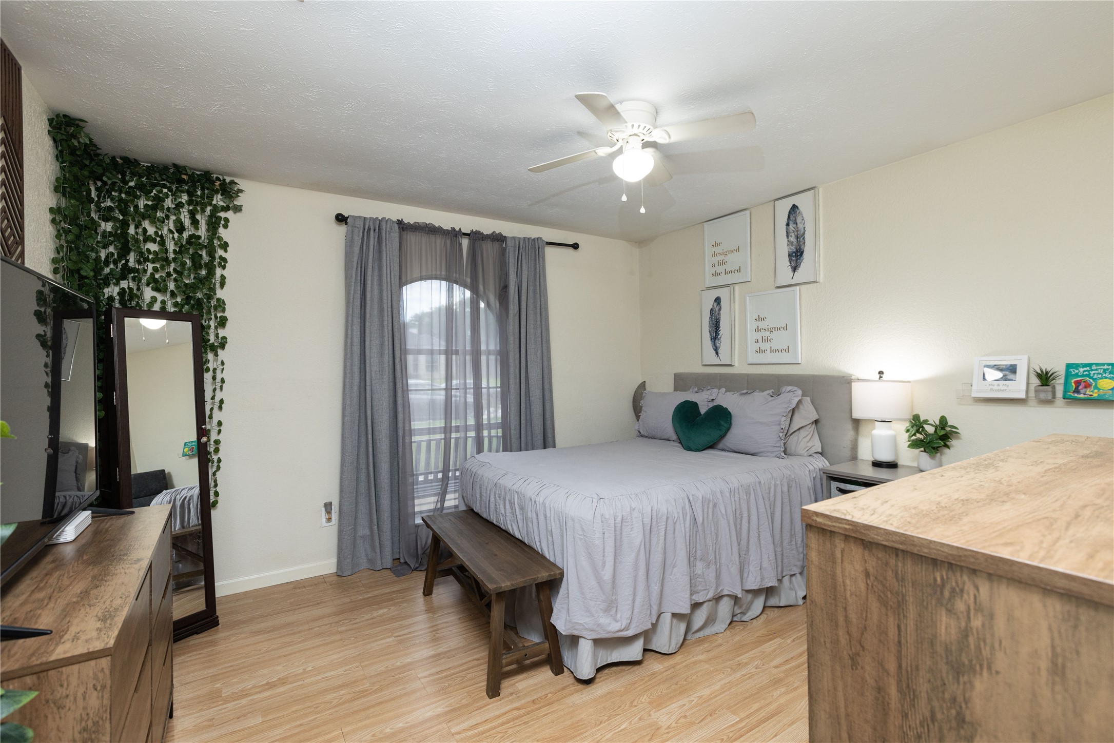 Cozy bedroom with a calming neutral palette features a comfortable bed, elegant wall art, and a ceiling fan for added comfort. Natural light filters through sheer curtains, highlighting the warm wood flooring and inviting decor.