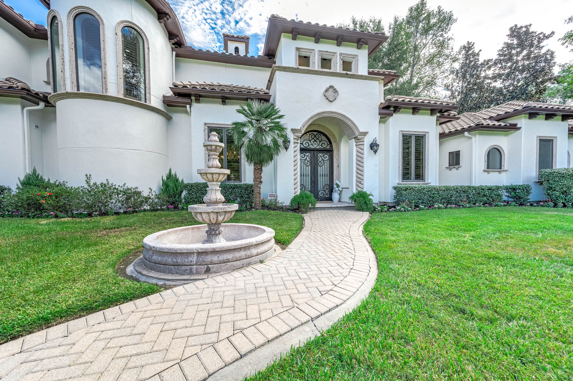 A paver walkway leads past a tiered stone fountain to the arched entry with wrought iron double doors.