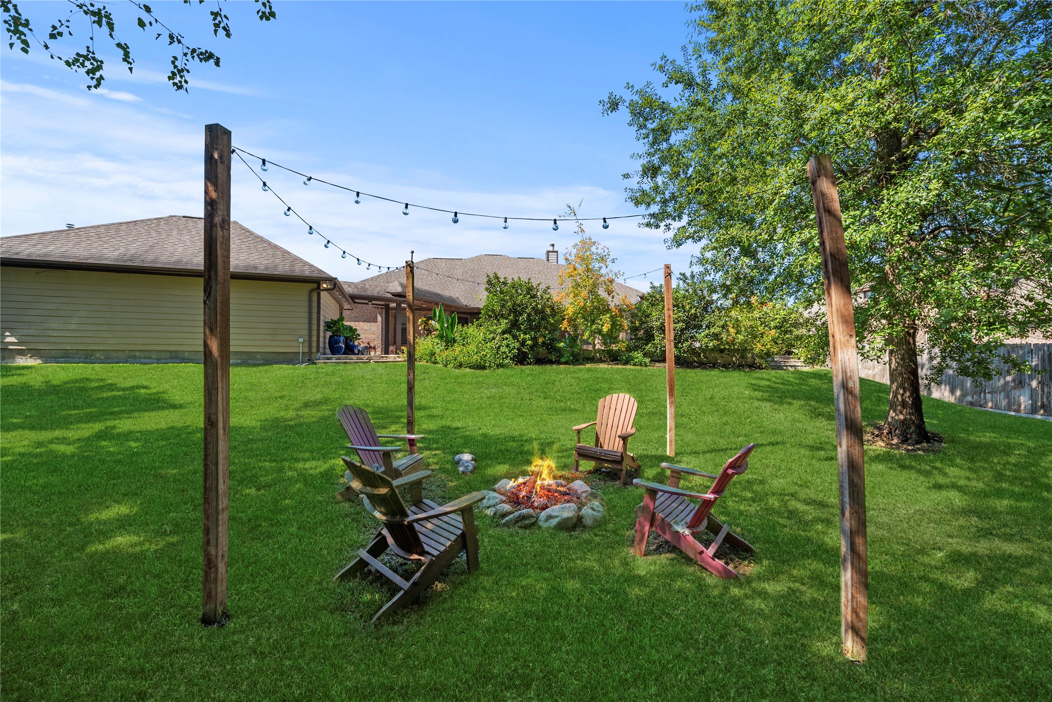 The perfect gathering spot featuring a charming fire pit surrounded by overhead string lighting, a lush green lawn, and mature shade trees.