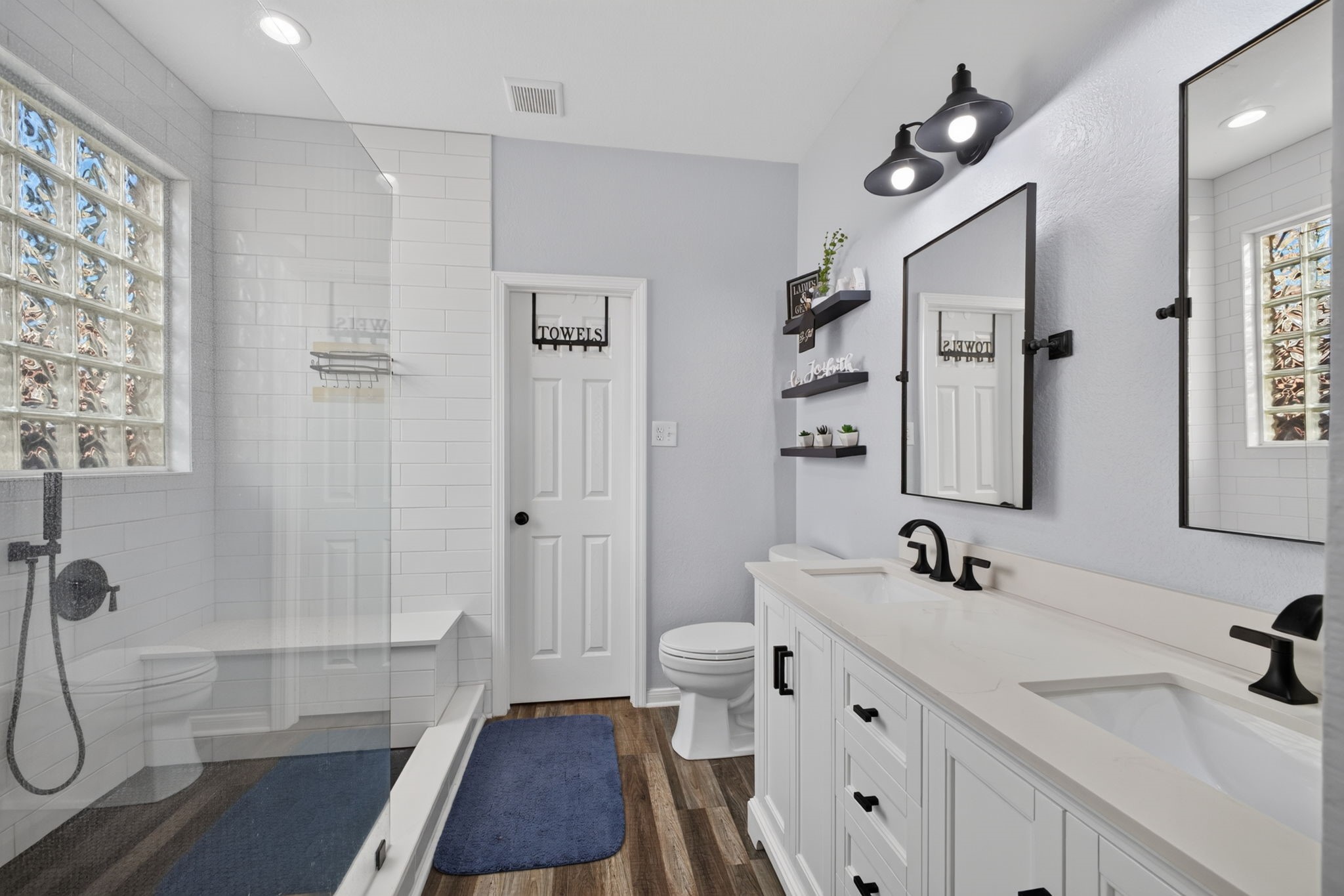 Modern bathroom with a sleek double vanity and black fixtures. Features a spacious glass-enclosed shower, subway tile, and natural light from glass block windows.