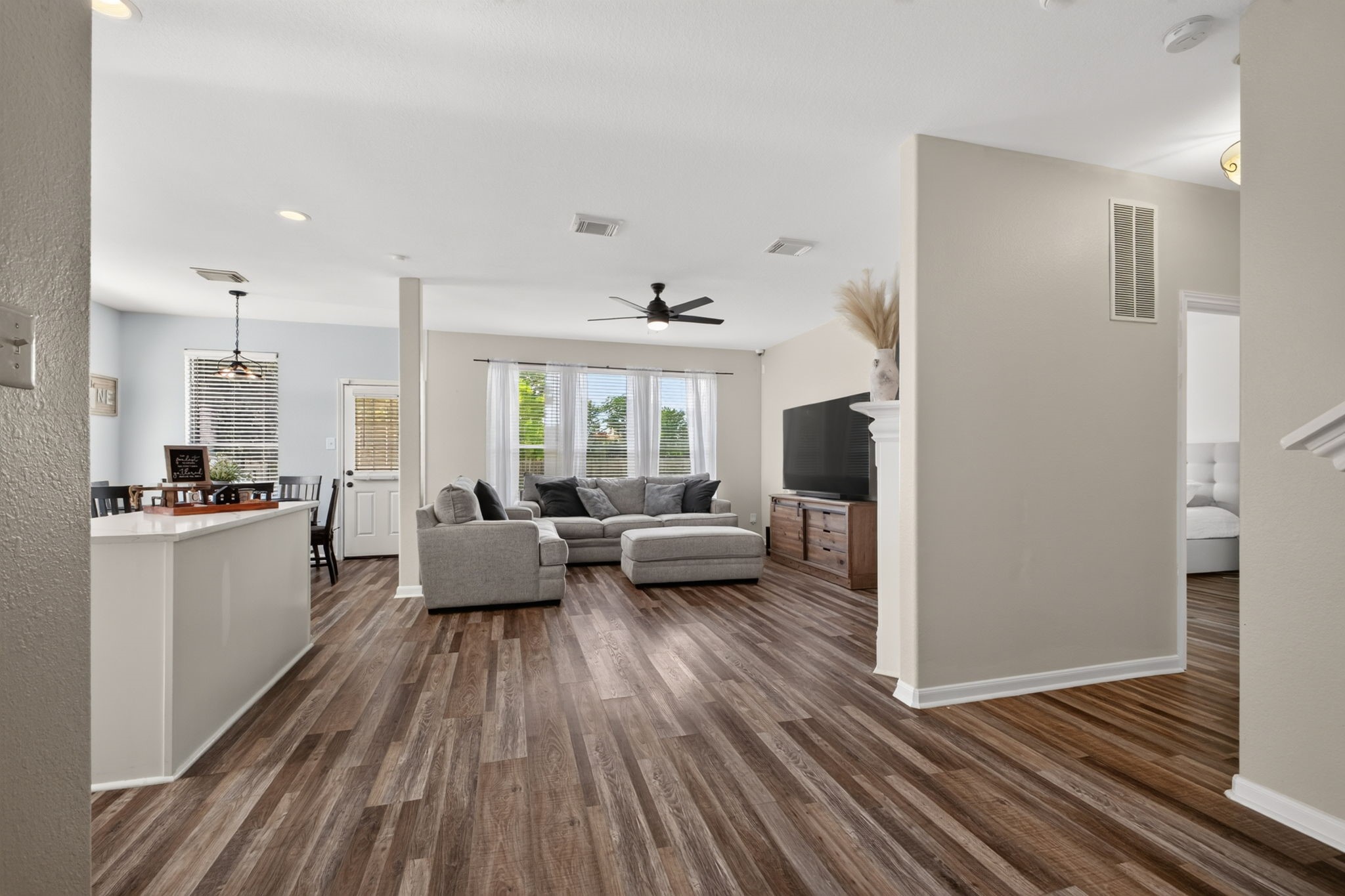 This spacious living area features an open floor plan with stylish flooring, abundant natural light from large windows, and a modern ceiling fan. The layout seamlessly connects the living room to a cozy dining area and kitchen, offering a welcoming space for relaxation and entertaining.
