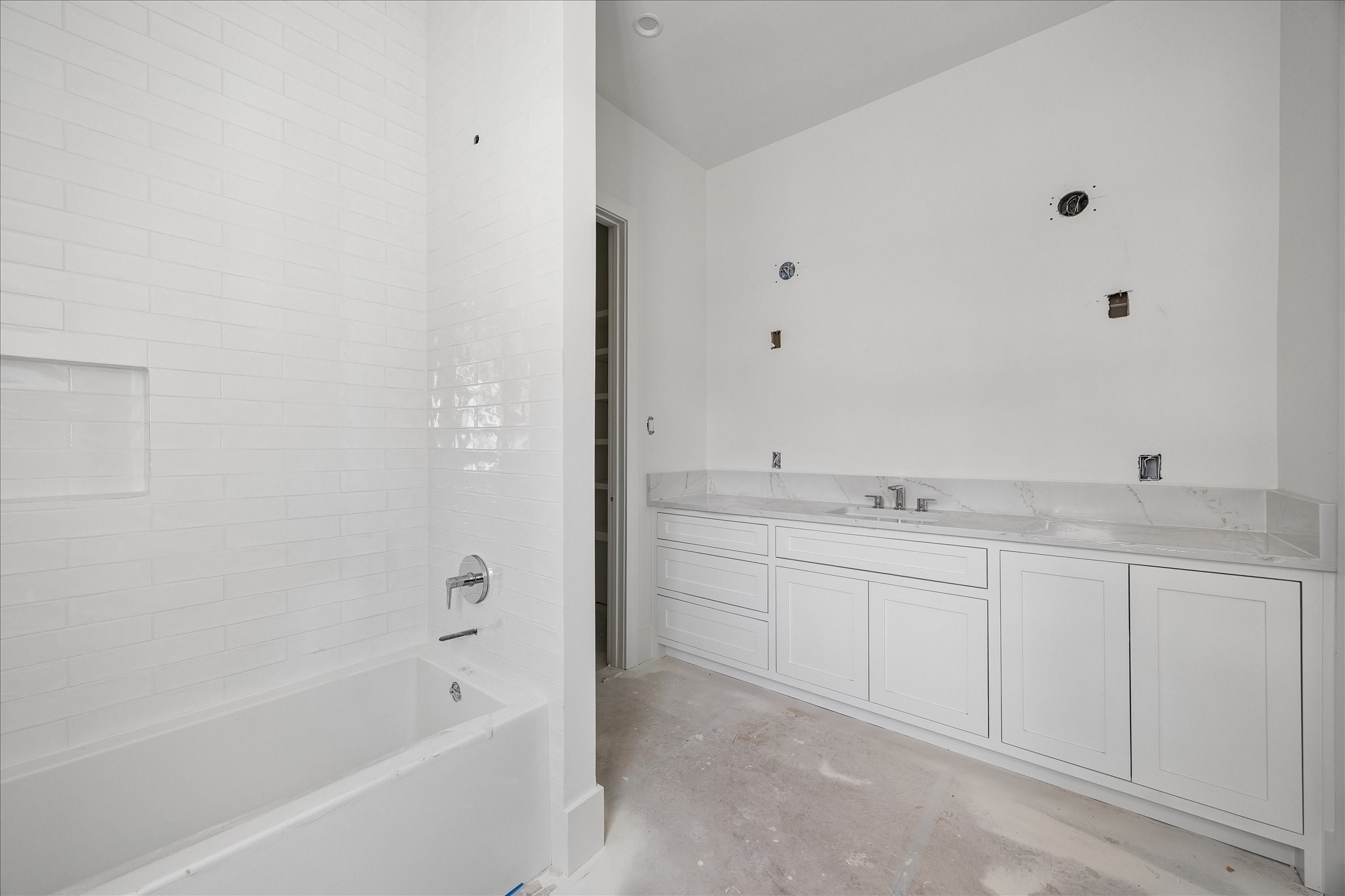 Wide vanity cabinetry extends the counter around a single basin, combining drawer storage with enclosed cabinets beside the tub. White tile continues up the bathing wall and around a recessed niche. Mirror, lighting and final trim remain in progress.