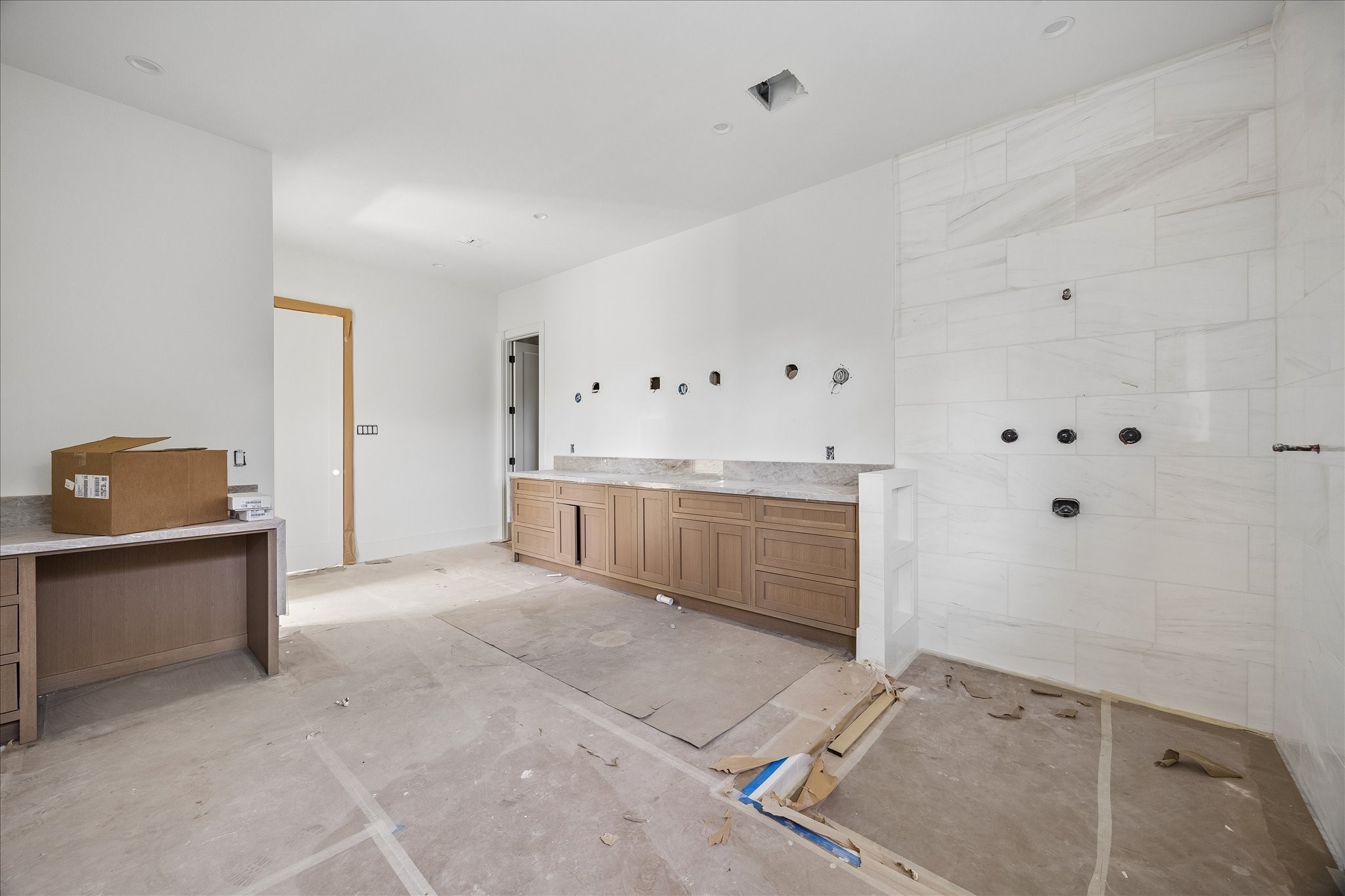 Long vanity cabinetry combines drawers and enclosed storage beside a broad tiled bathing area. Natural tone wood and light veined tile establish the palette, while wall plumbing rough ins and protective floor coverings show the bath before fixtures and final fittings are installed.