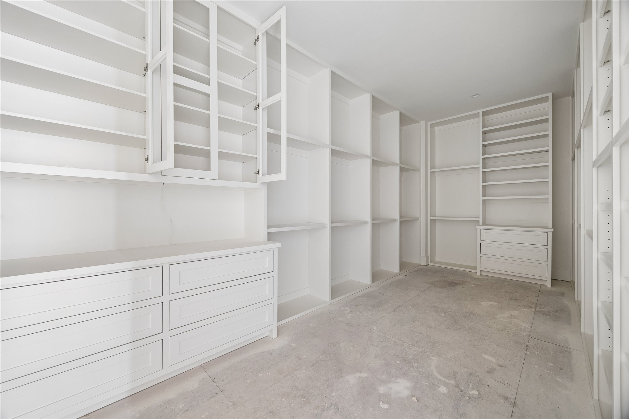 Custom closet cabinetry combines open shelves, drawer banks and tall compartments for different storage needs. Storage extends around the perimeter and uses the full wall height, leaving a central passage through the room. Flooring and final hardware remain in progress.