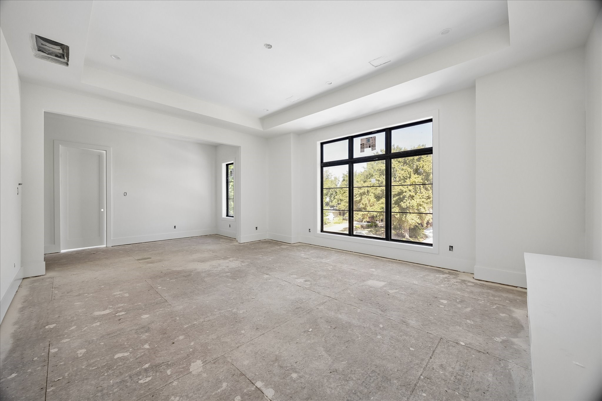 Large windows and a recessed ceiling give this upper level room a bright, open character. A wall recess adds definition beside the doorway without closing the room off. The view shows the room before flooring and lighting installation are complete.