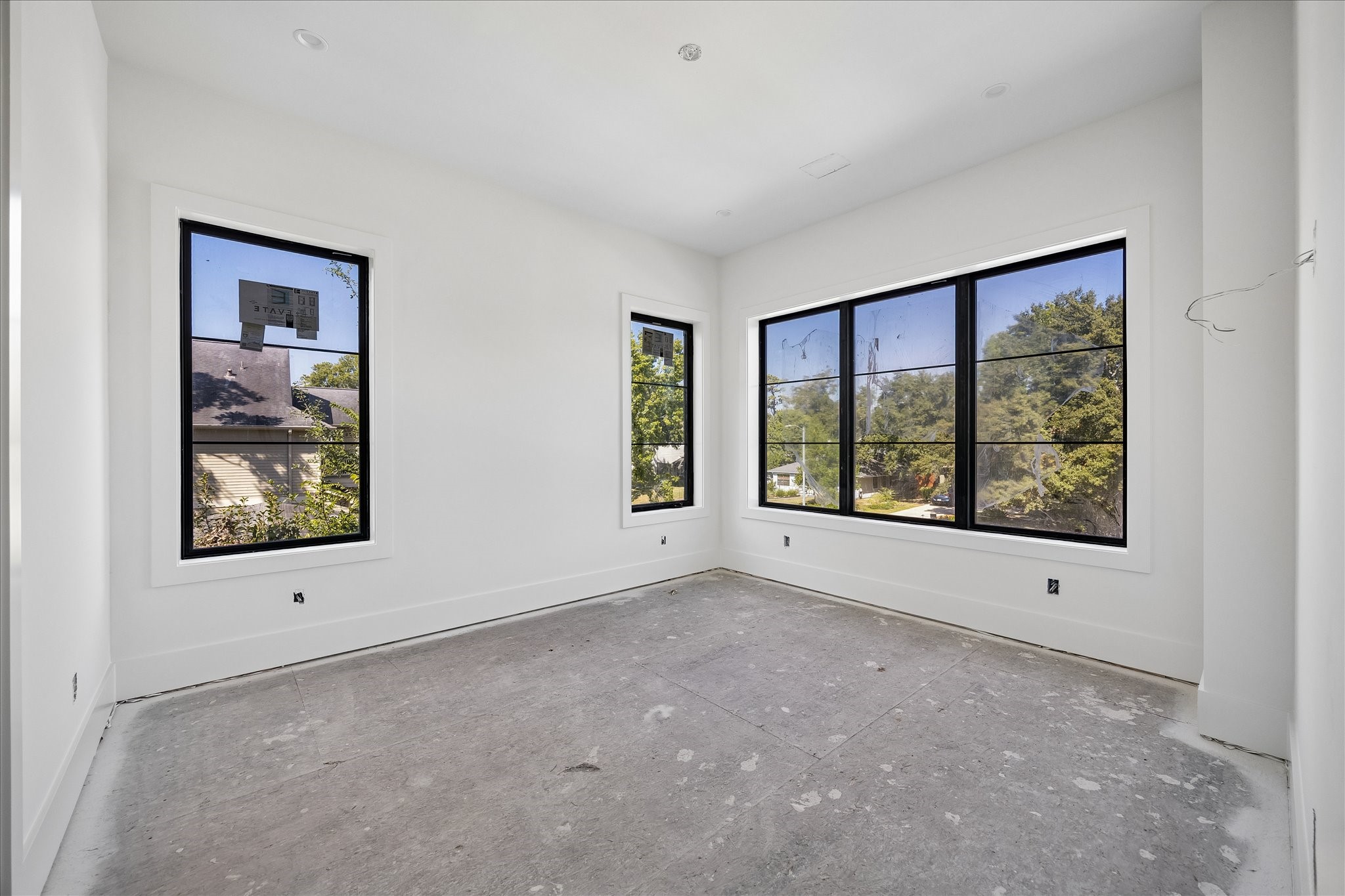 Windows on 2 walls bring daylight from different directions, including a broad view toward the trees outside. Black frames contrast with the light wall and trim palette, while the uninterrupted wall areas leave room for the space's intended use. Final flooring is still underway.