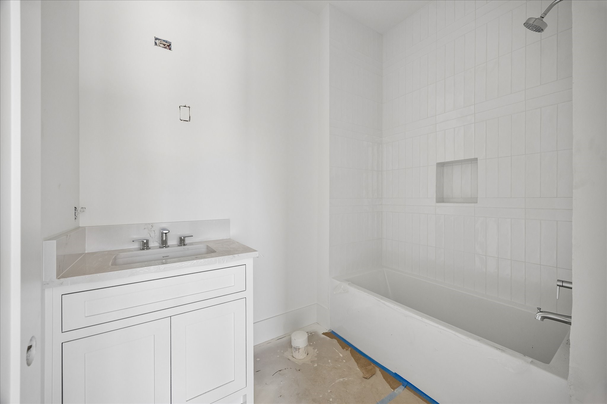 Single basin vanity storage sits beside a tub with a tiled shower surround and recessed niche. The light counter and white cabinetry keep the bath palette restrained, with chrome fittings adding a reflective accent. Mirror, lighting and final trim remain in progress.