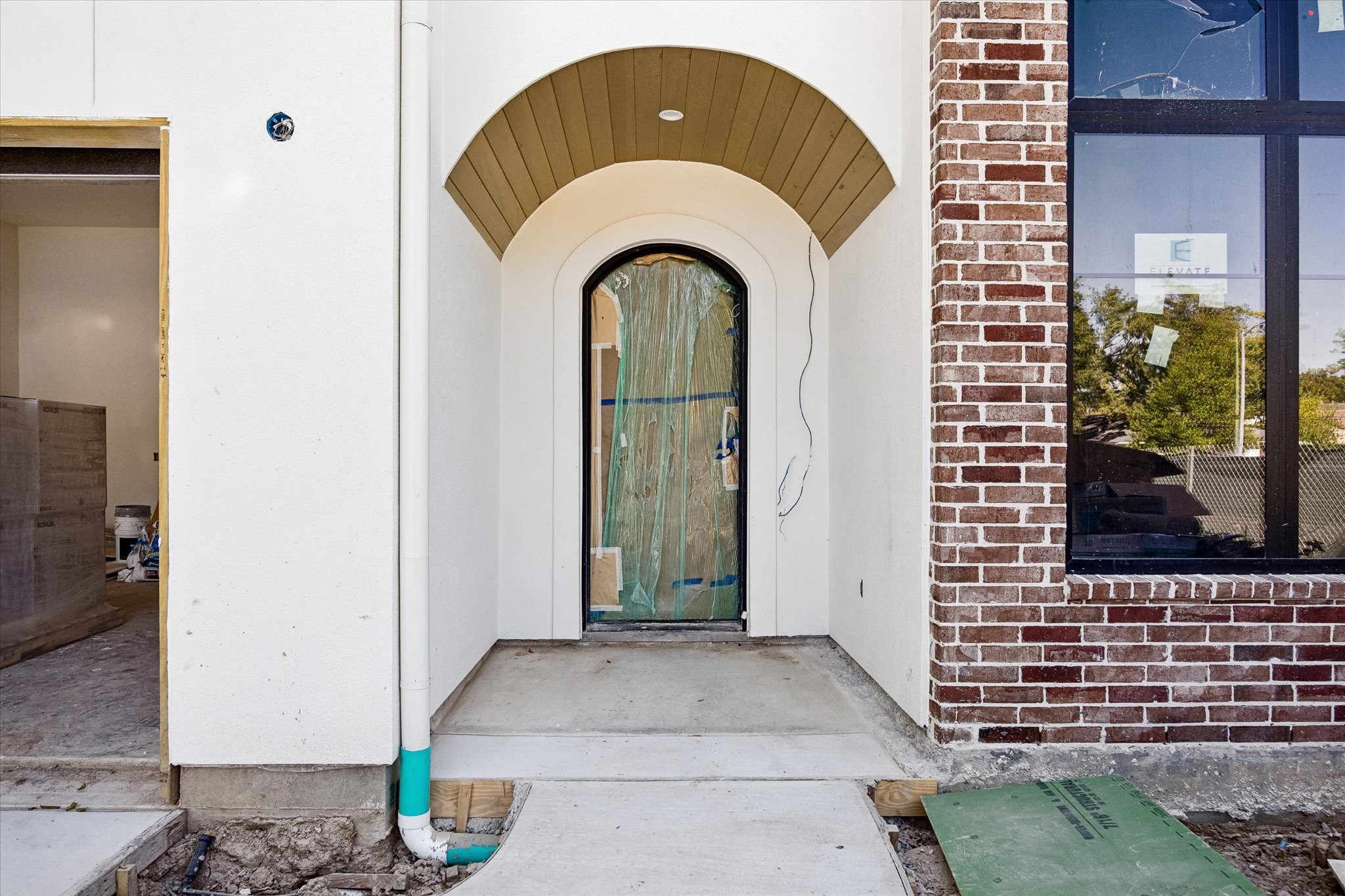 Recessed arches shelter the front door beneath a stained beadboard porch ceiling. Brick texture, a tall black framed window and the curved entry opening layer the exterior materials at the approach. Protective covering remains on the door during construction.