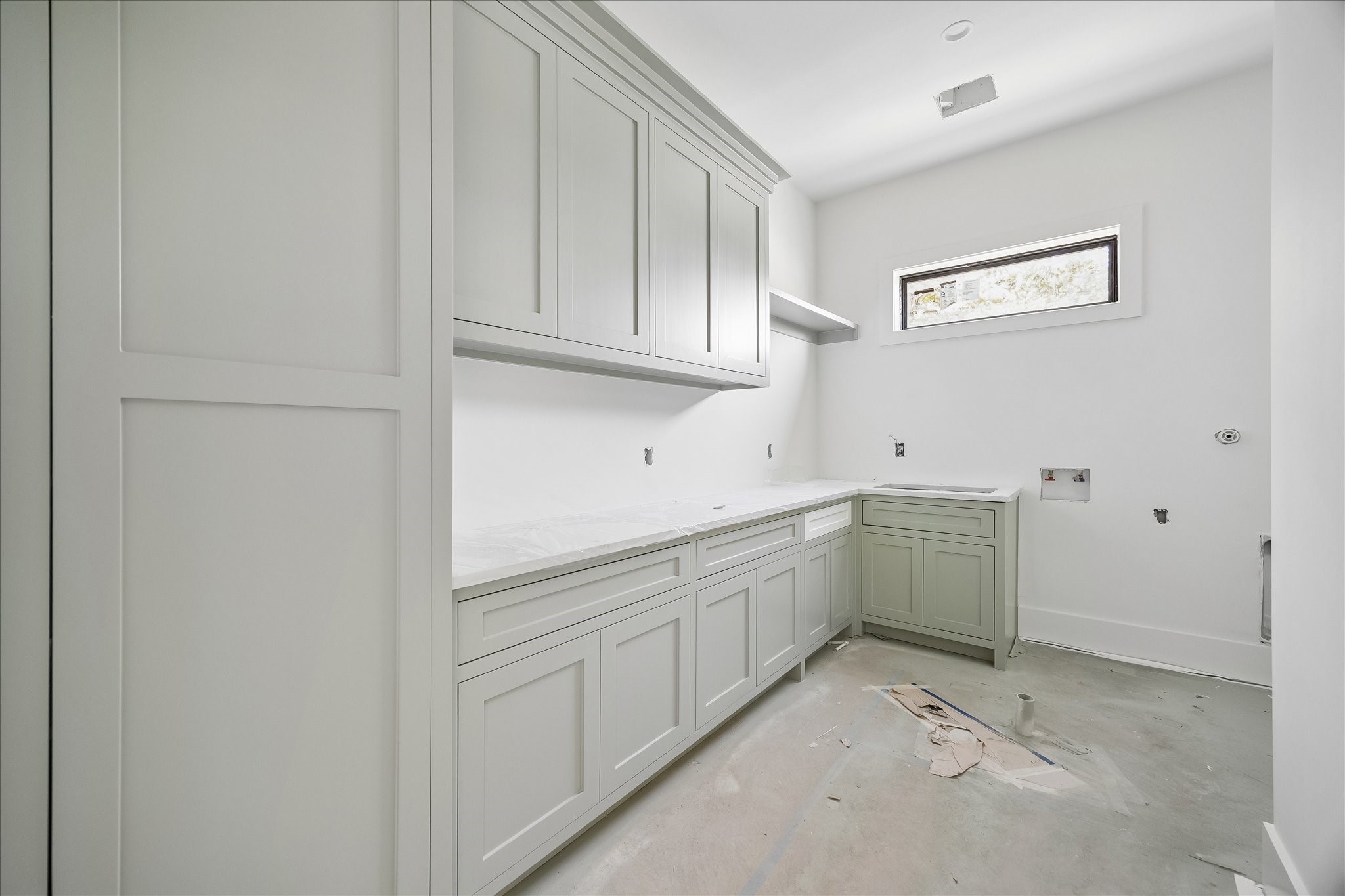 Laundry counters and cabinetry return around the corner, with a sink beneath the horizontal window and a drying rod below the short shelf. Upper cabinets add storage. The builder also confirmed an outlet inside the vacuum storage cabinet for concealed charging; appliances are not installed.
