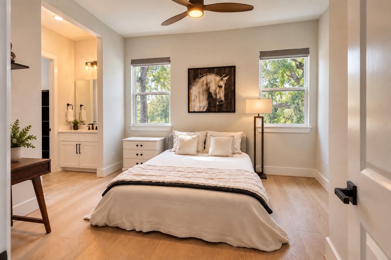 Bright and inviting bedroom featuring large windows with serene tree views, light wood flooring, a modern ceiling fan, and an ensuite vanity area.