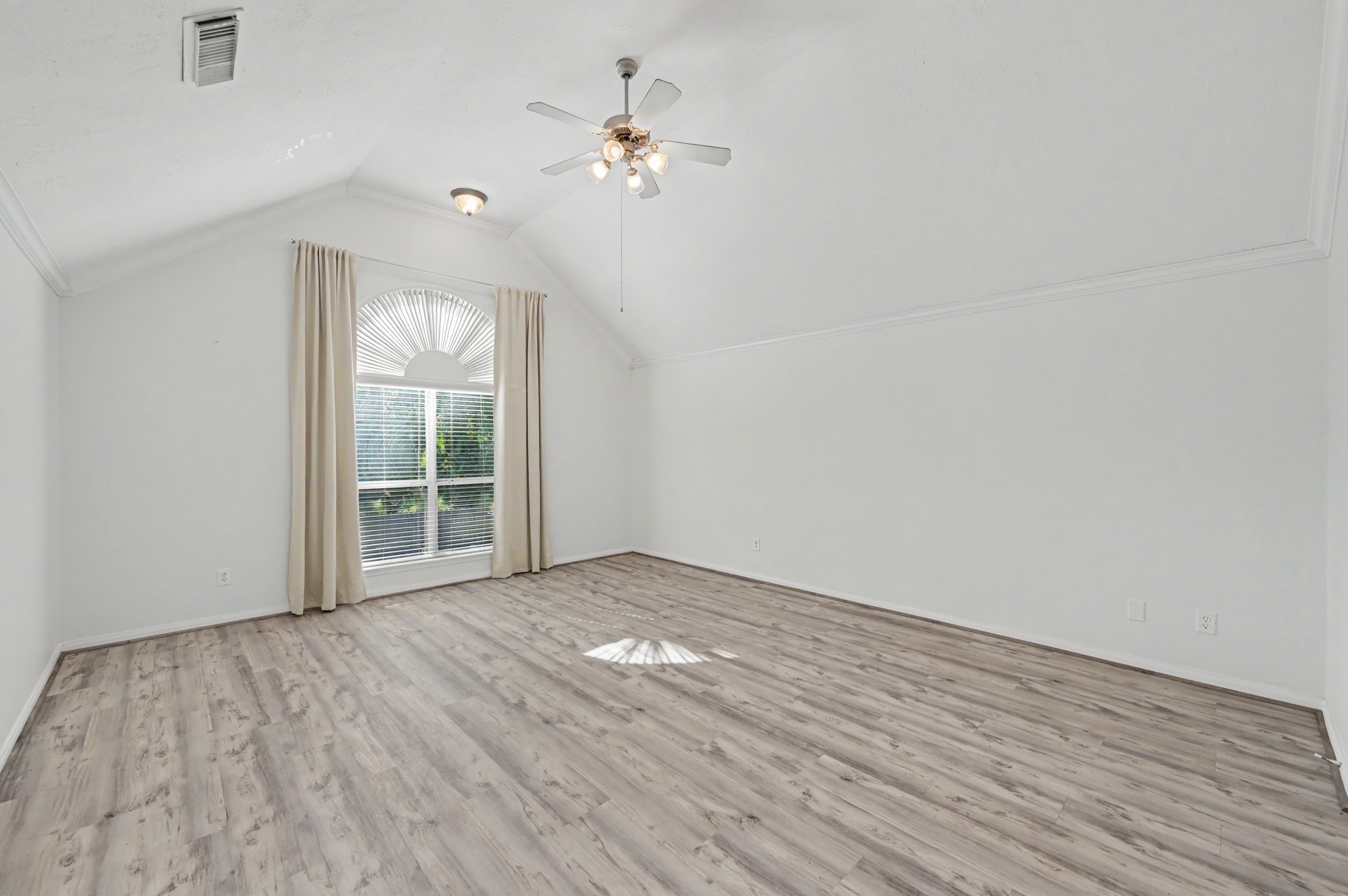 Oversized primary suite featuring vaulted ceilings, crown molding, wood-look flooring, ceiling fan and large arched window with abundant natural light.