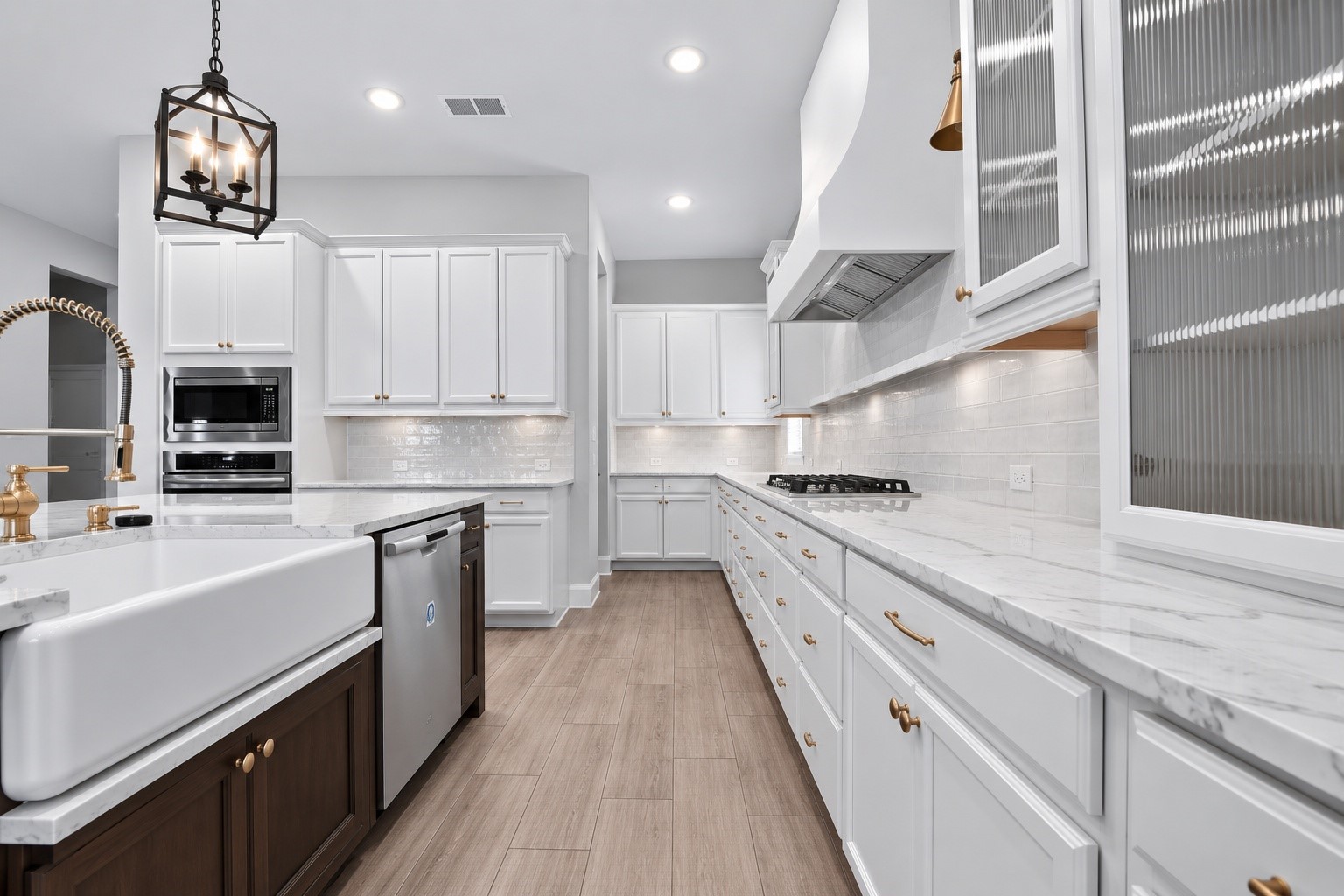 Spacious gourmet kitchen featuring white cabinetry, quartz countertops, a large contrasting island, farmhouse sink, stainless steel appliances, and beautiful gold accents throughout.