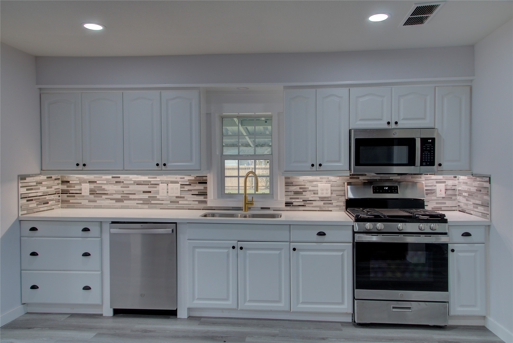 This modern kitchen features sleek white cabinetry, stainless steel appliances, and a stylish tile backsplash. It includes a gas stove, microwave, dishwasher, and a window above the sink, providing ample natural light.