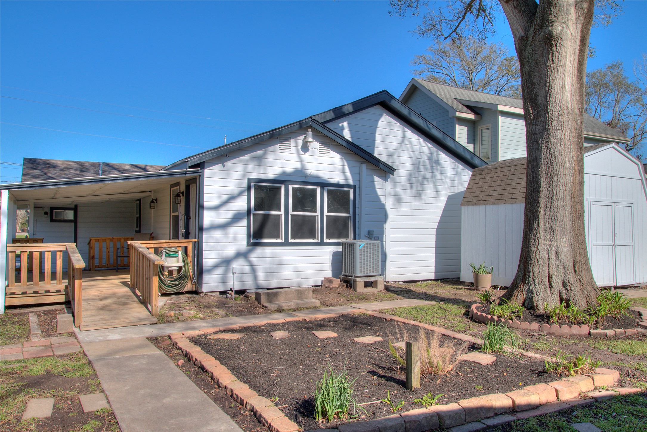 Charming covered patio with a wooden deck, convenient ramps, and a mature shade tree overlooking a cozy garden area.