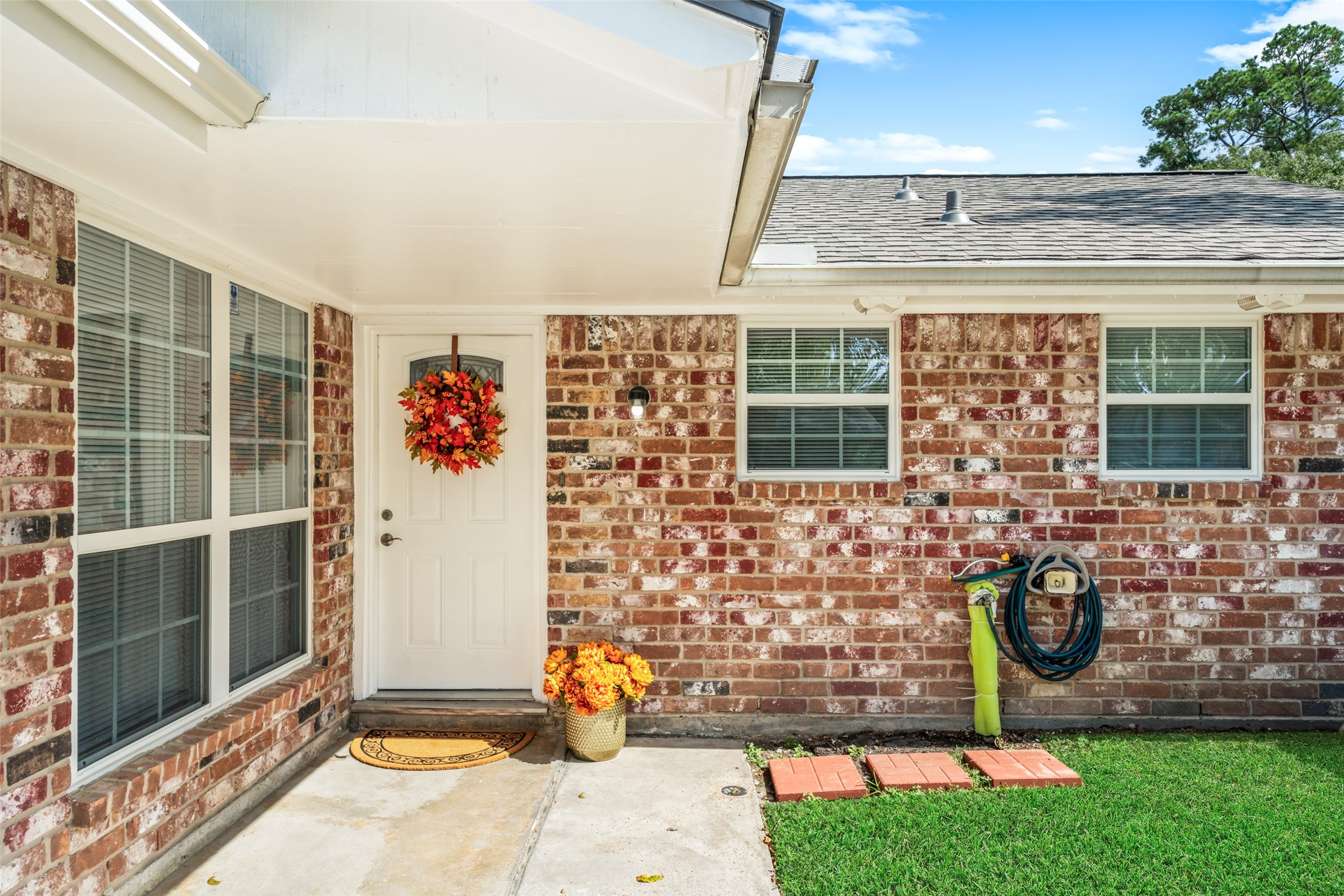 Covered entryway features classic brickwork, large windows, a clean concrete walkway, and a neatly maintained lawn. The outside white wood areas of the home have been freshly painted, giving the exterior a crisp, refreshed appearance.