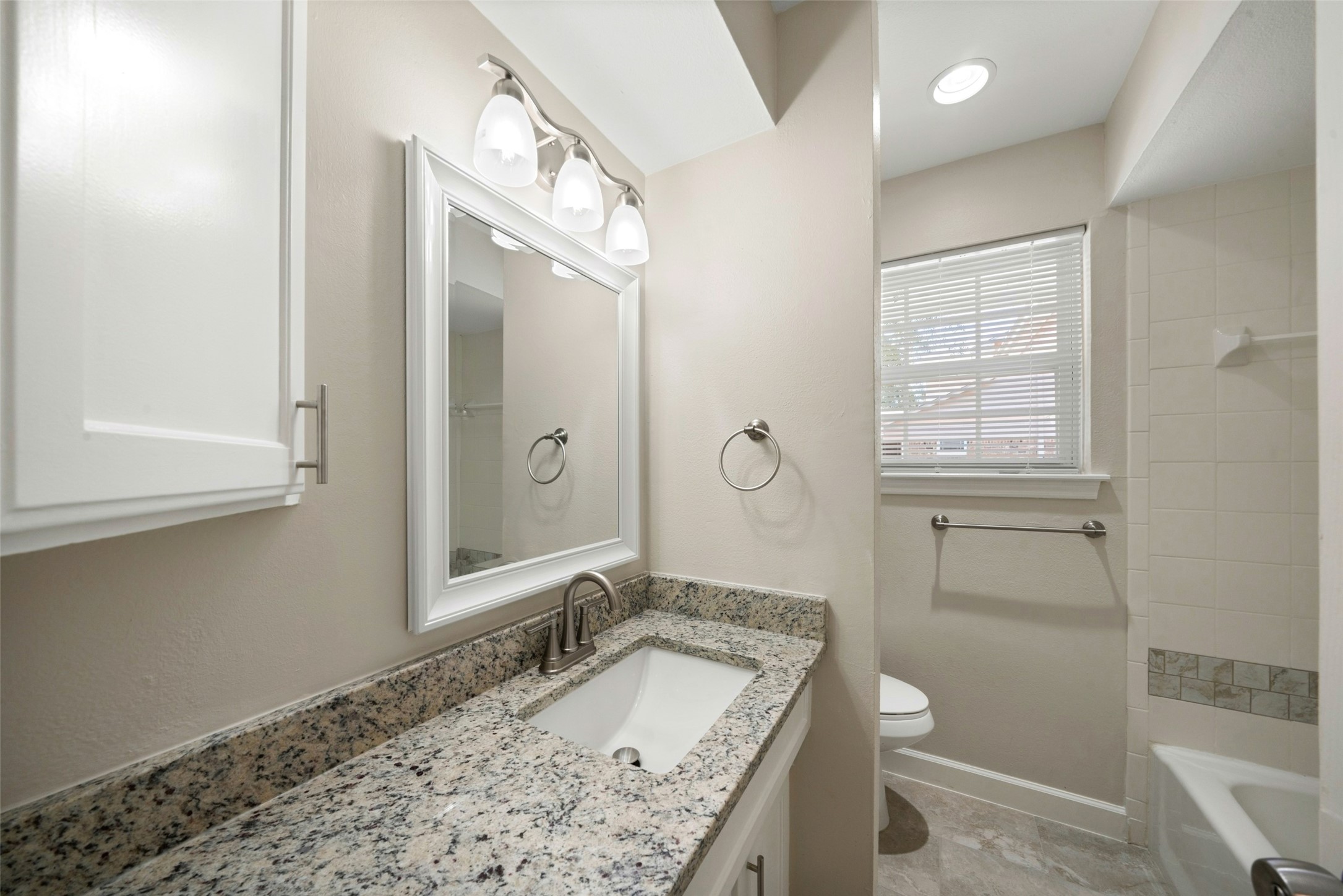This bright, updated hall bathroom features a clean neutral color palette, a new granite countertop, an undermount sink with brushed nickel faucet, and a crisp white-framed mirror that adds a fresh, polished look. The space is finished with coordinating brushed nickel fixtures, ample lighting, and a tiled tub/shower area for a clean and timeless feel.