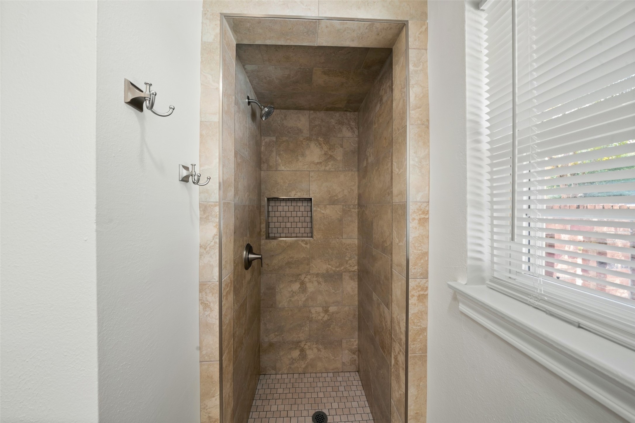 This updated primary bathroom features a newly tiled walk-in shower with warm neutral tones, a built-in recessed niche for added convenience, and coordinating tile flooring for a clean, finished look. The shower is complemented by brushed nickel faucets and fixtures, creating a polished, modern feel while maintaining a warm and timeless design.