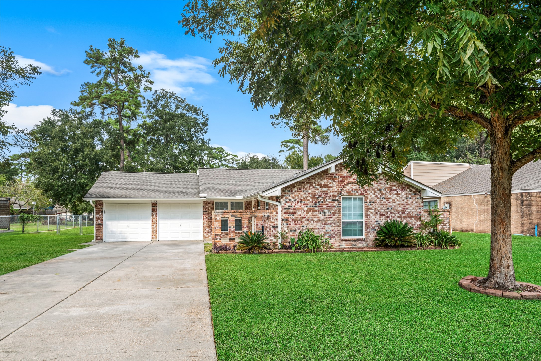 A charming single-story brick home featuring a two-car garage, spacious driveway, and a beautifully maintained green lawn. Mature trees provide plenty of shade, while simple landscaping around the front adds a welcoming touch to the homes classic exterior.
