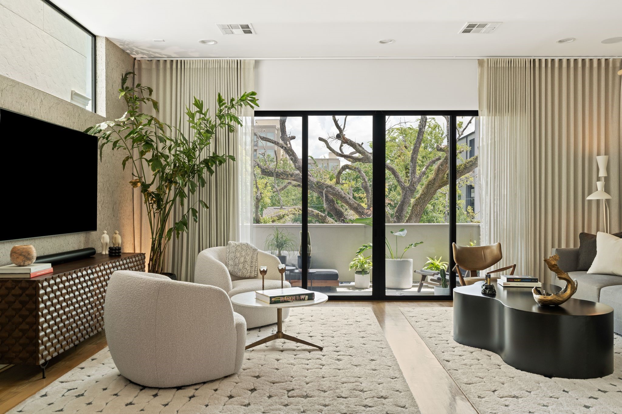 Light-filled second-floor living room with hardwood floors, professionally curated interiors, custom drapery and expansive sliding glass doors opening to the covered terrace and mature oak canopy. Light-filled second-floor living room with hardwood floors, professionally curated interiors, custom drapery and expansive sliding glass doors opening to the covered terrace and mature oak canopy.