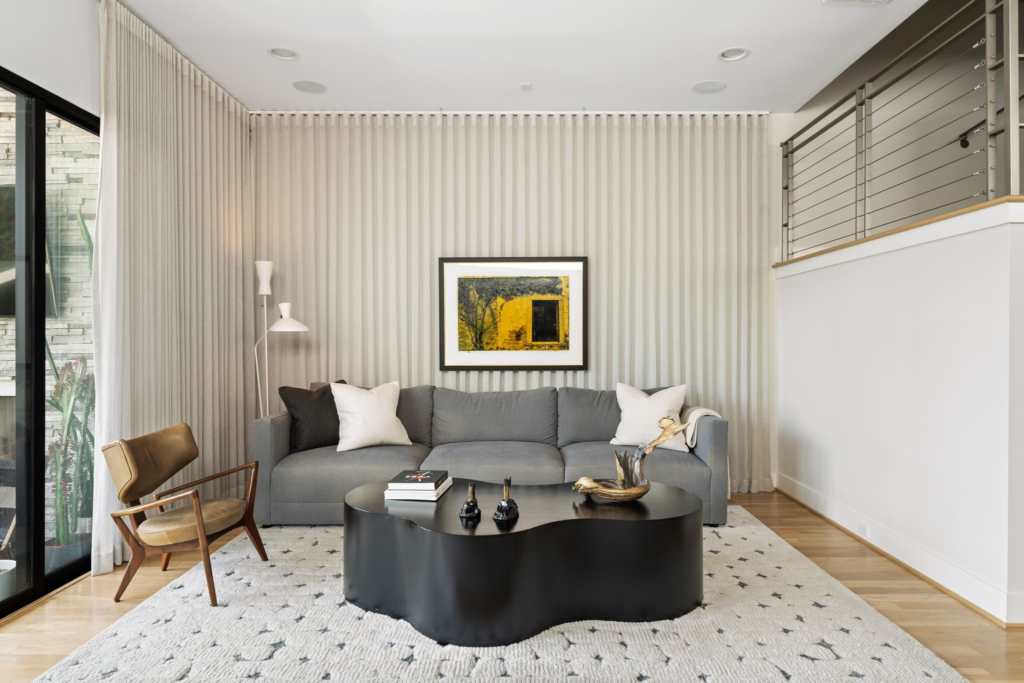 Professionally curated living room features dramatic custom drapery extending across the wall, ceiling-suspended artwork, hardwood floors, and a sophisticated layered aesthetic.