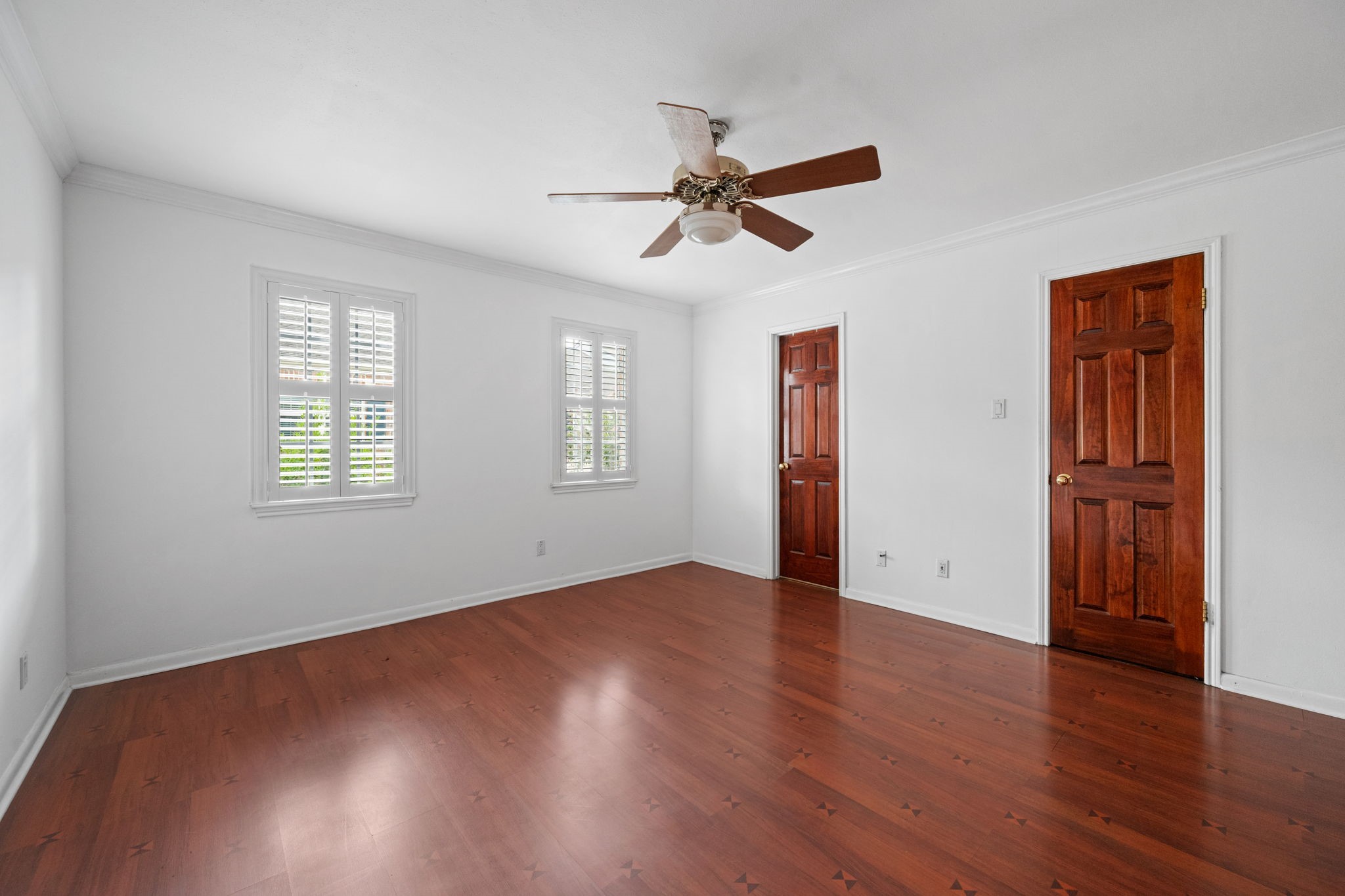 Bright and spacious room featuring rich hardwood floors, two wooden doors, and large windows with shutters, complemented by a ceiling fan.