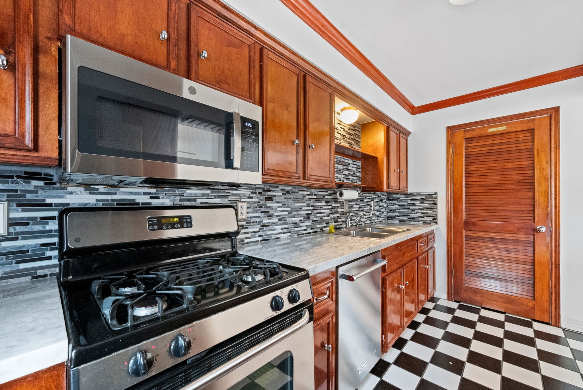 Stylish kitchen featuring modern stainless steel appliances, a sleek tile backsplash, rich wood cabinetry, and classic black-and-white checkered flooring for a timeless appeal.