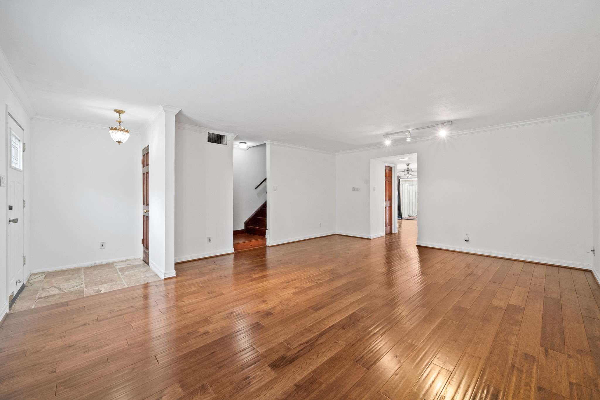 Spacious living area with elegant hardwood floors and bright white walls, featuring recessed lighting and a charming chandelier, leading to a staircase and additional rooms. Spacious living area with elegant hardwood floors and bright white walls, featuring recessed lighting and a charming chandelier, leading to a staircase and additional rooms.
