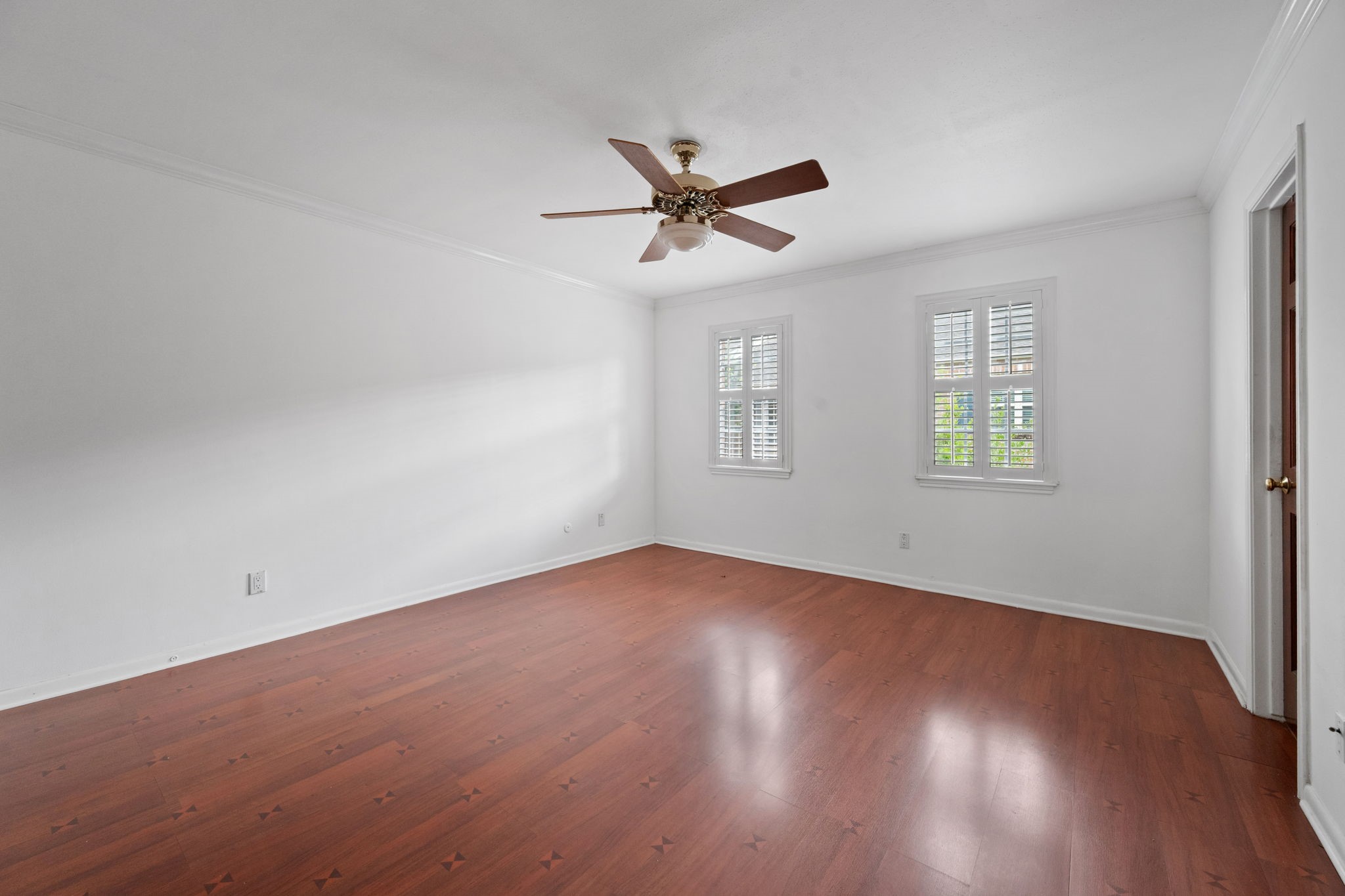 Spacious bedroom with rich hardwood floors, elegant white walls, and abundant natural light from dual windows with plantation shutters. Features a ceiling fan for added comfort.