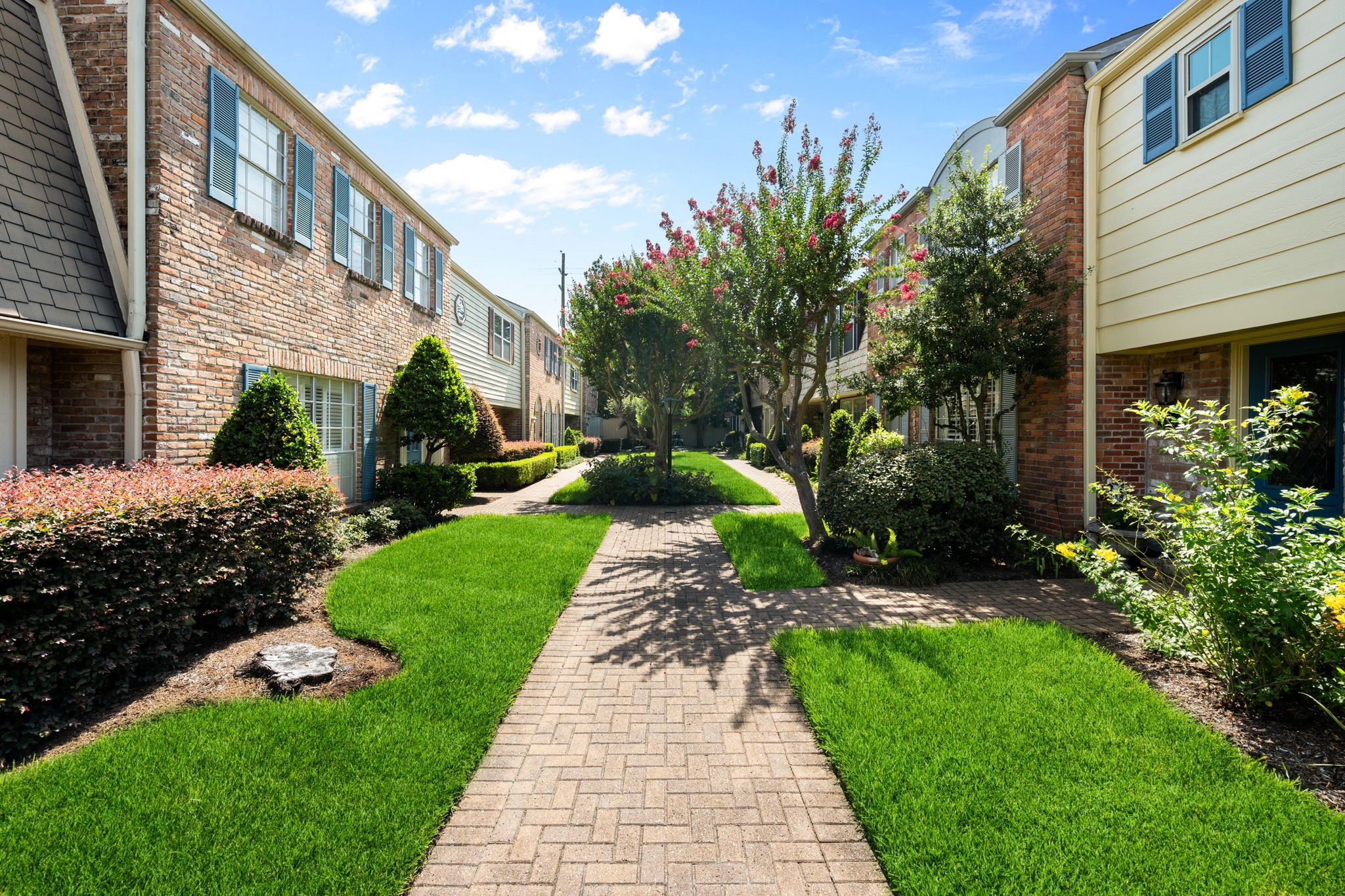 Charming townhomes with classic brick facades line a beautifully landscaped walkway, featuring lush greenery and mature trees under a bright blue sky.