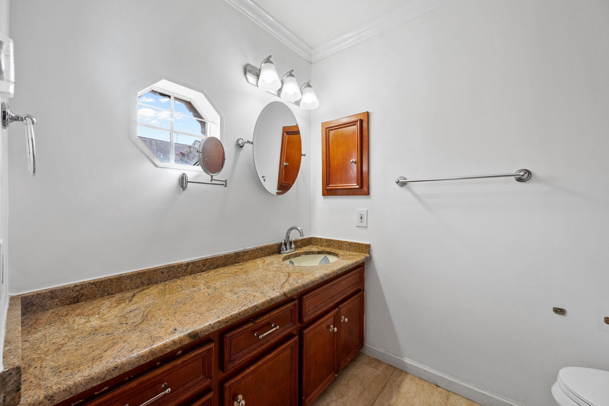 Elegant bathroom featuring a spacious granite countertop with rich wood cabinetry, complemented by a stylish octagonal window and modern lighting fixtures.