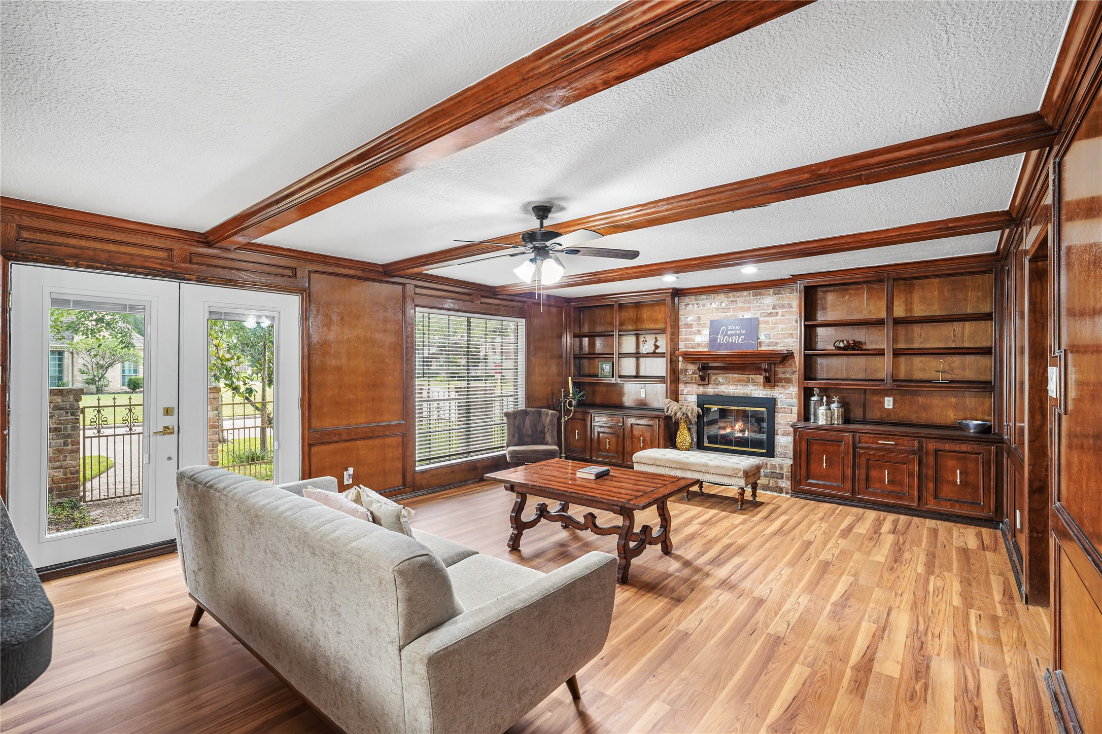Elegant living room featuring rich wood paneling, a cozy fireplace, built-in shelving, and French doors opening to a serene outdoor view.