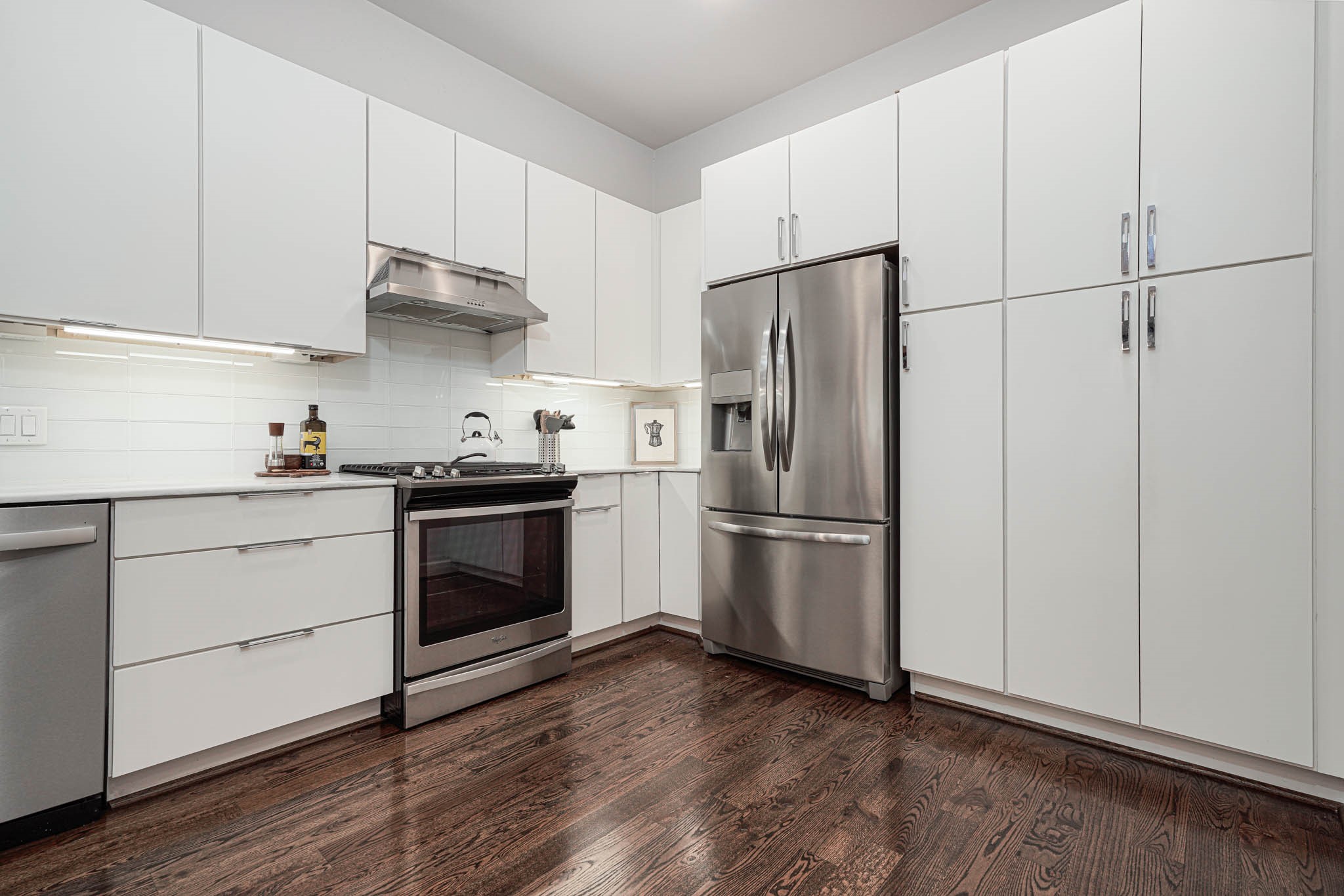 A modern kitchen features sleek white cabinetry, stainless steel appliances, and rich hardwood floors, creating a stylish and functional space.