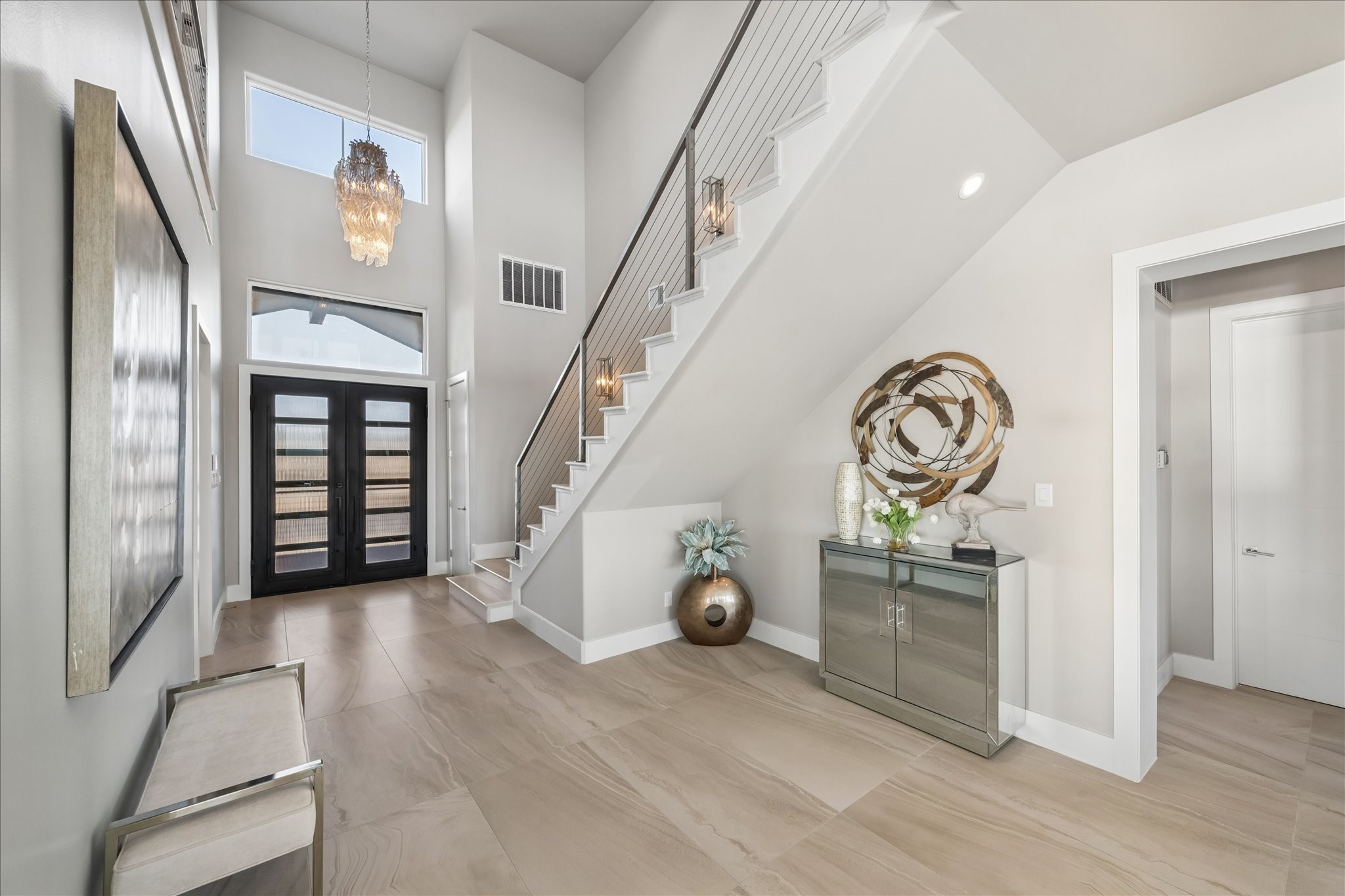 A grand, foyer showcases soaring ceilings, abundant natural light, and modern double doors with glass lites and a transom window. A staircase with wood treads and cable railing anchors the space, complemented by a dramatic chandelier and sleek Eleganza tile flooring