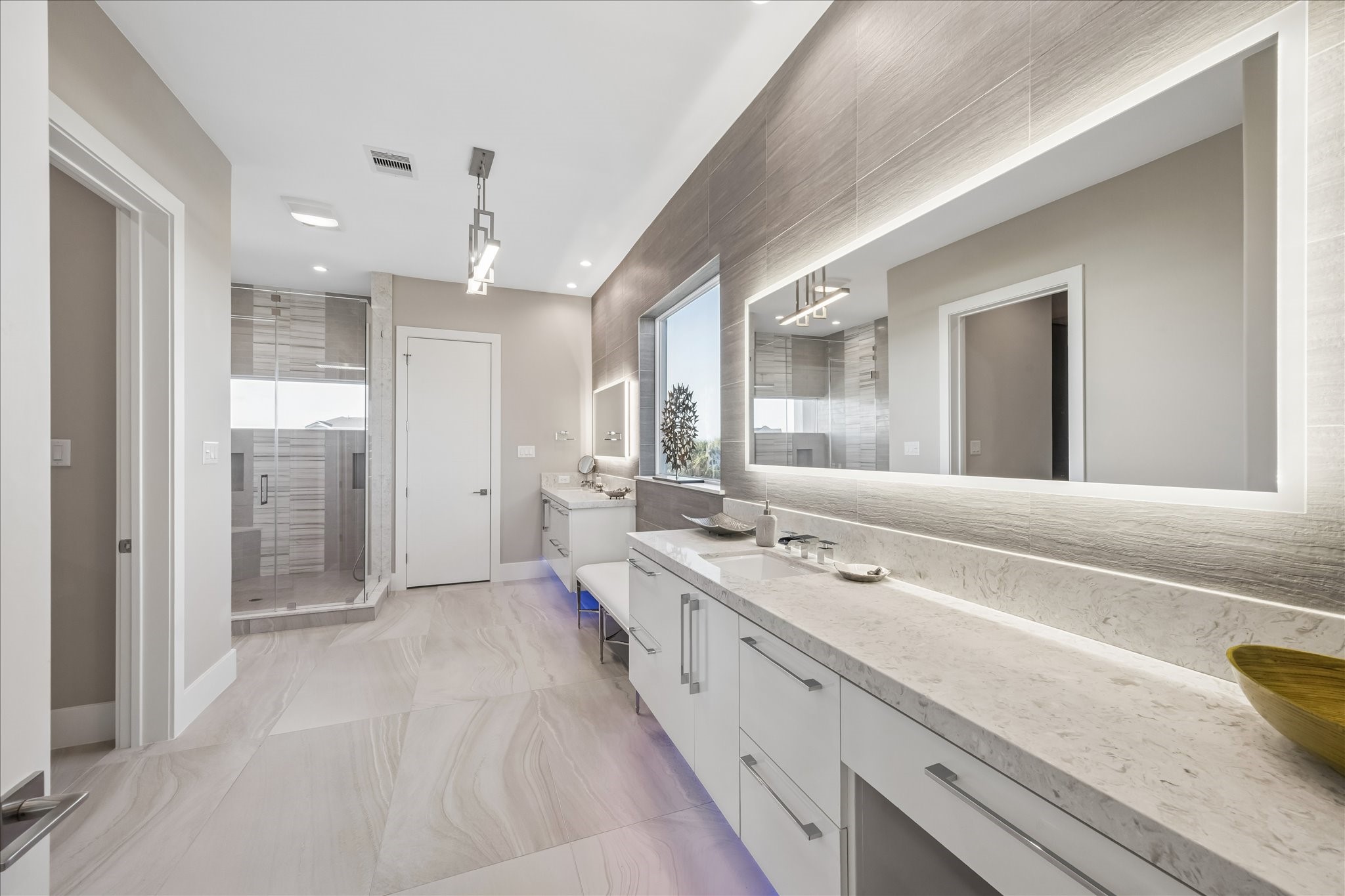 The primary ensuite bathroom is bright and modern, featuring an elongated double vanity with quartz countertops, large backlit mirrors, under-vanity lighting, a frameless walk-in shower, and a comfortable sitting area