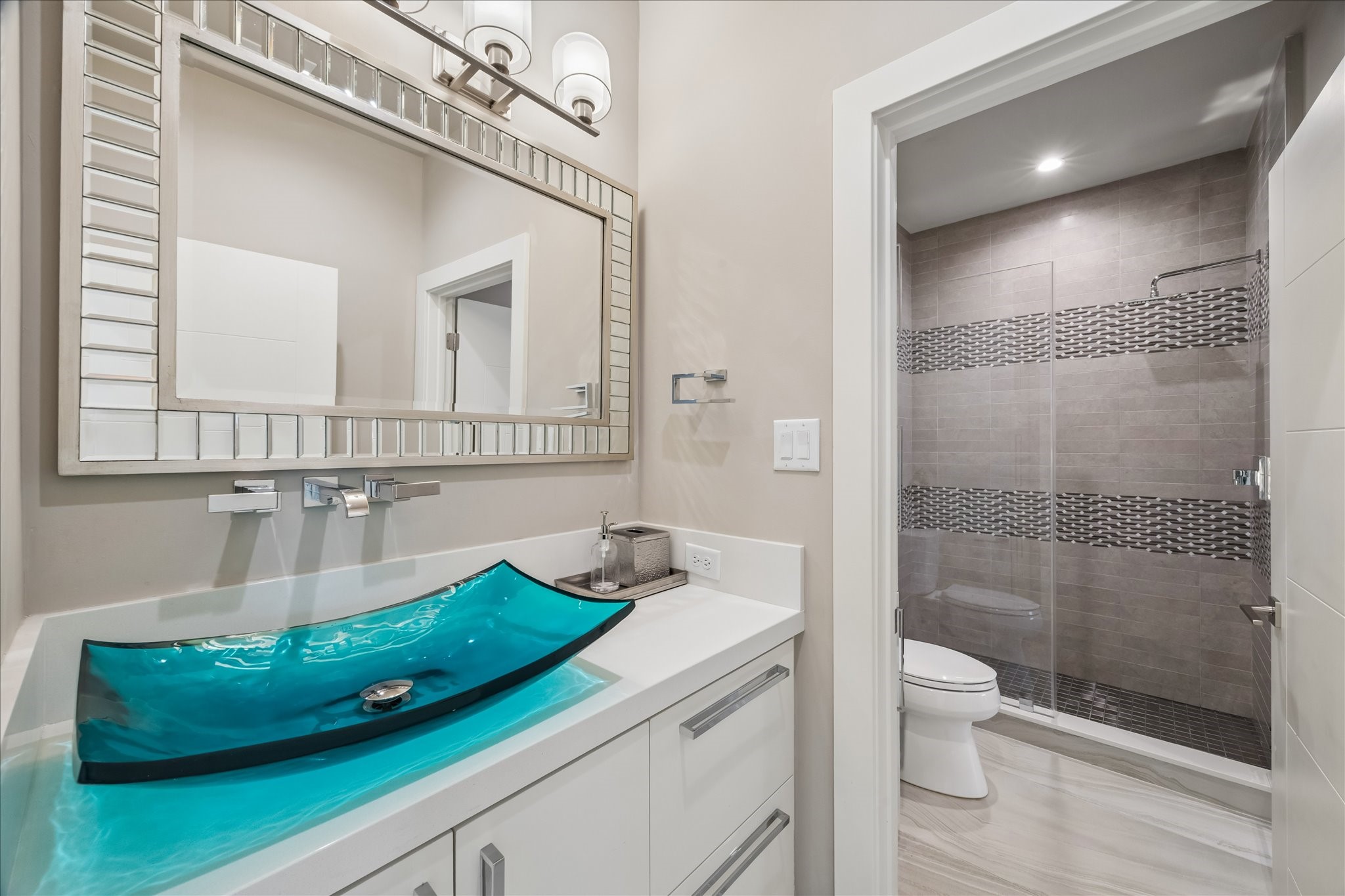 The guest bathroom showcases a clean, modern color palette with a double vanity setupone dedicated to the guest bedroom for privacy and an additional vanity accessible from the family room. Features include a turquoise glass vessel sink, quartz countertop, under-vanity lighting, and a spacious walk-in shower