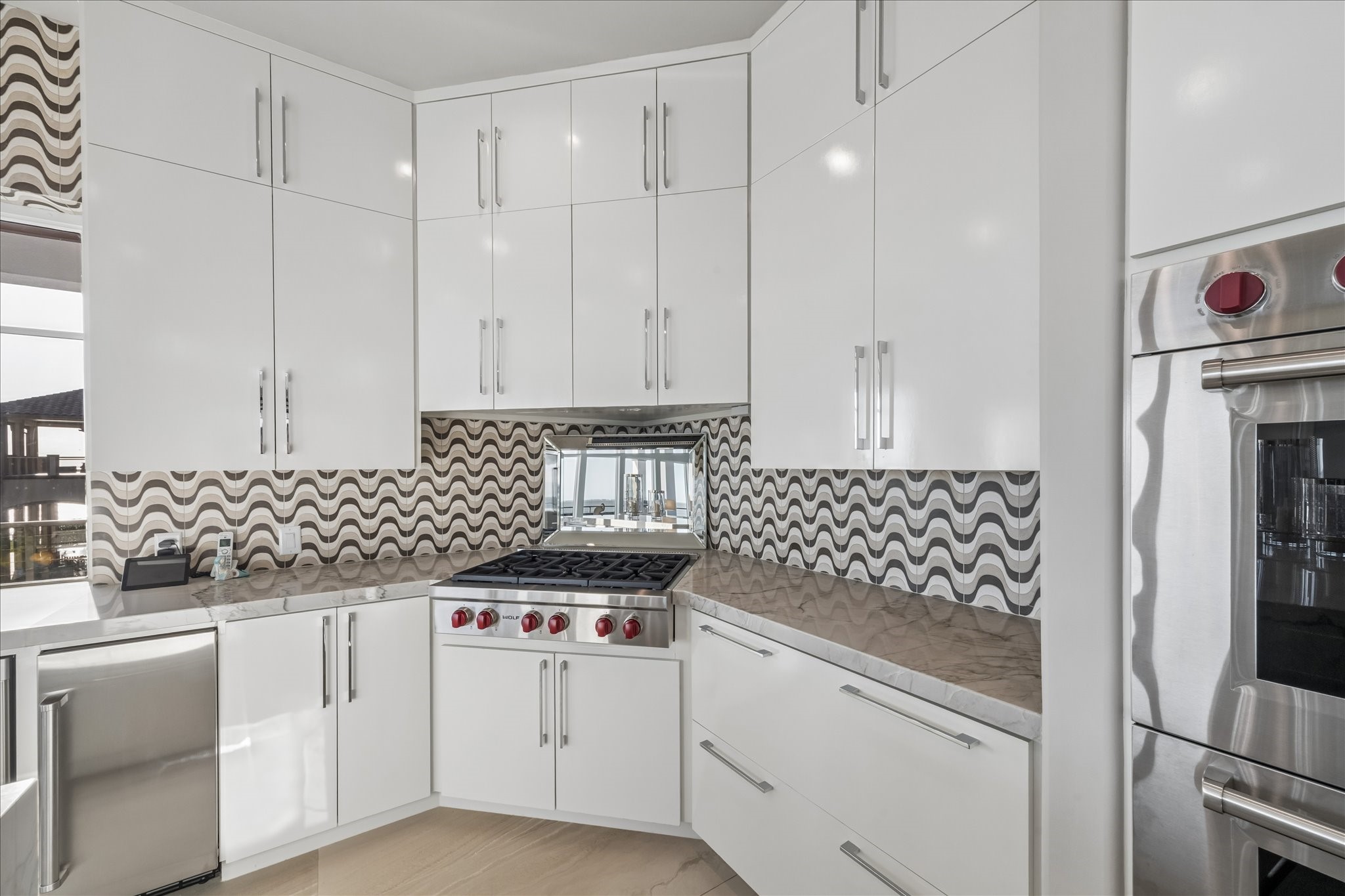 A mirrored accent behind the gas cooktop reflects both light and water views, amplifying the rooms bright, airy ambiance. The modern wavy tile backsplash brings depth and a contemporary style to the space